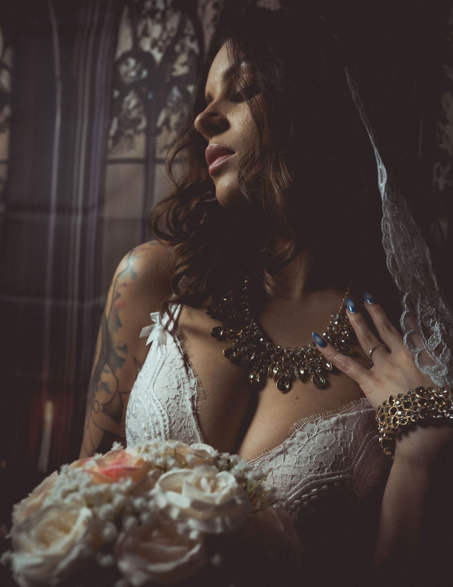 Tattooed bride in lace lingerie holding a floral bouquet, wearing a statement necklace and veil in moody lighting.
