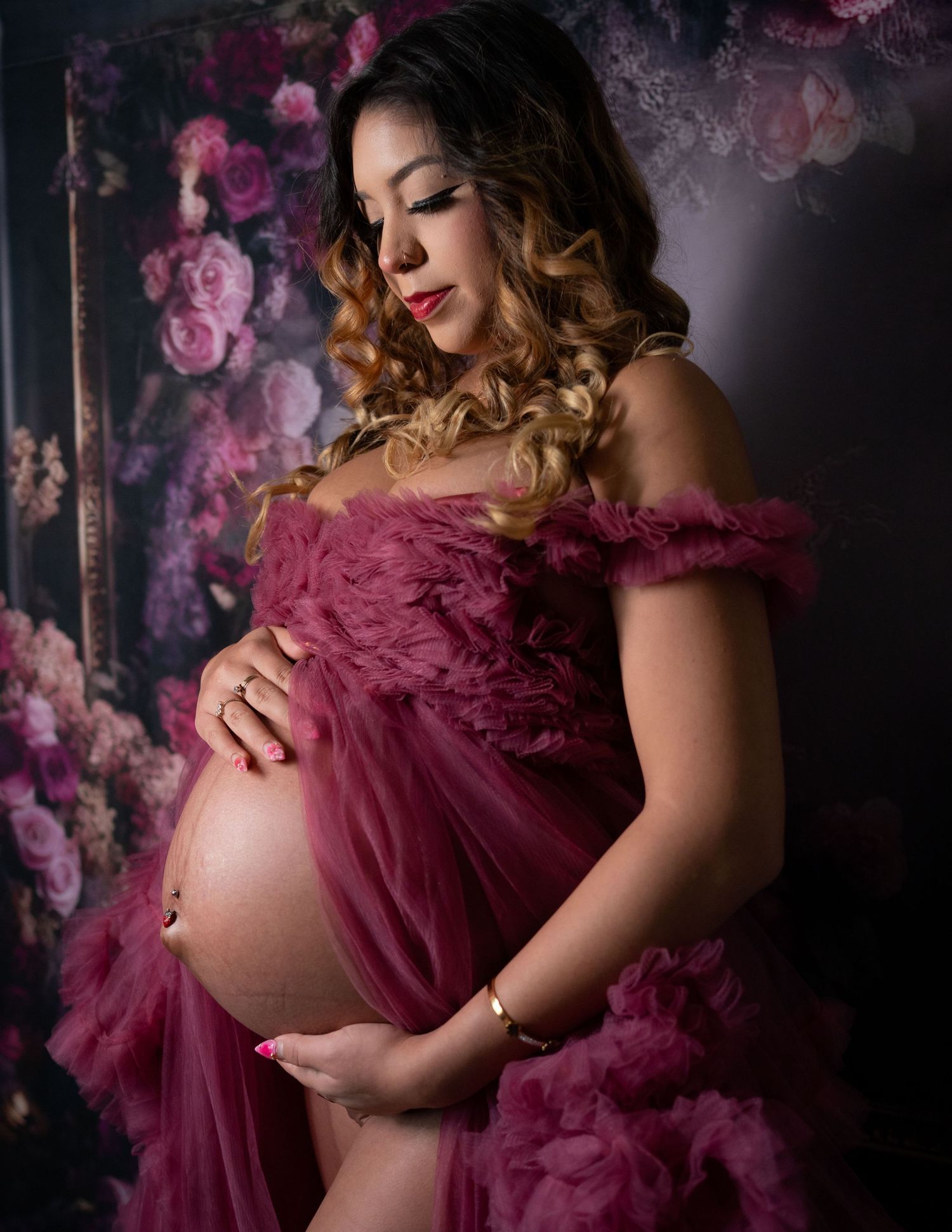 Pregnant woman in a burgundy tulle maternity gown cradling her baby bump against a floral backdrop.