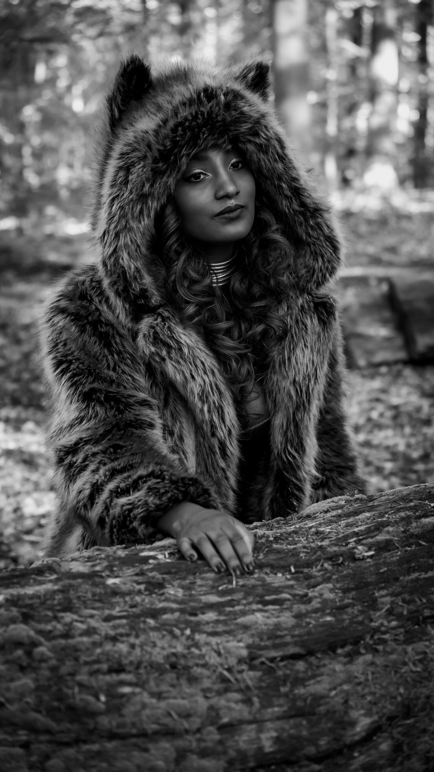A person wearing a luxurious furry hooded coat poses in a moody black and white forest setting.