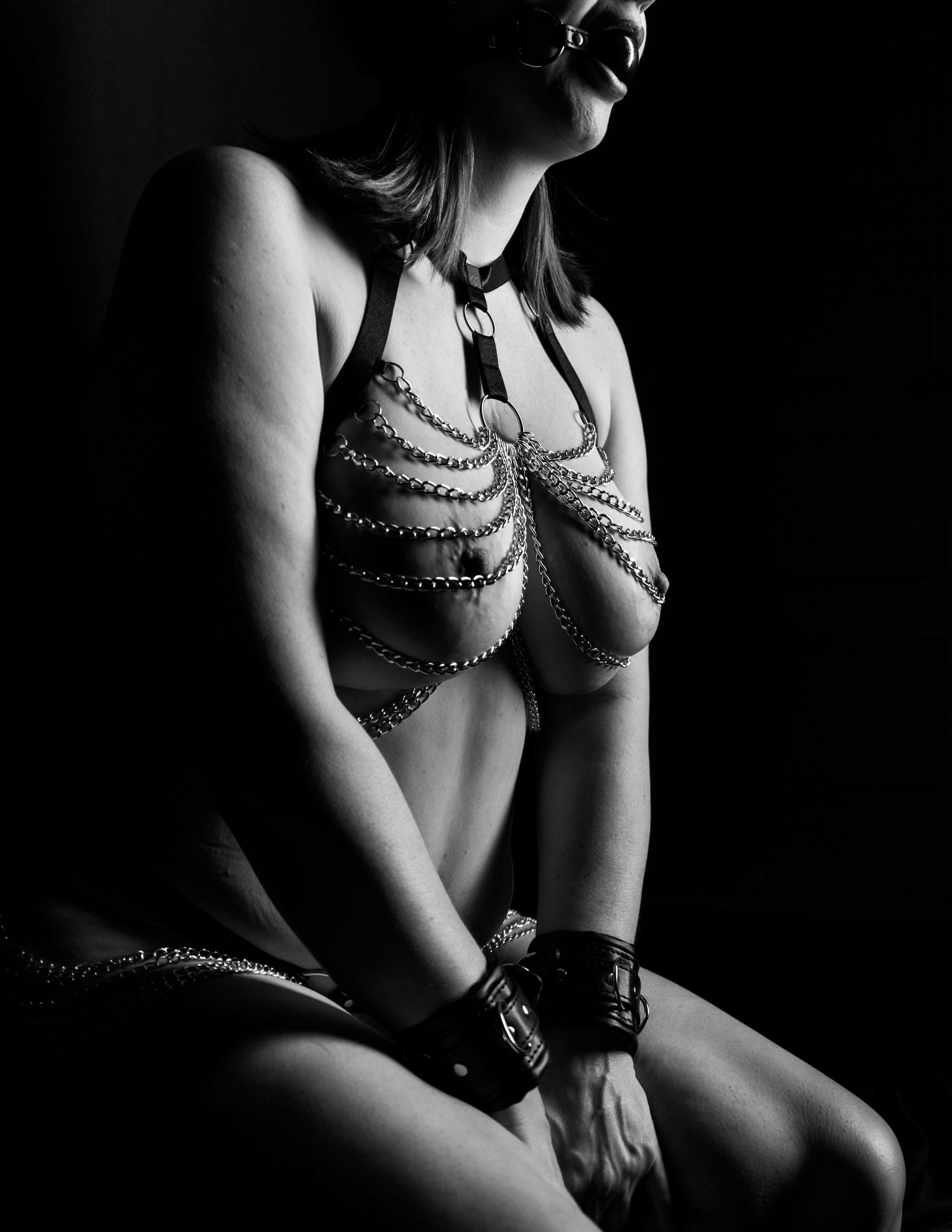 Black and white artistic photo of a woman wearing decorative body chains and leather wrist cuffs in dramatic lighting.