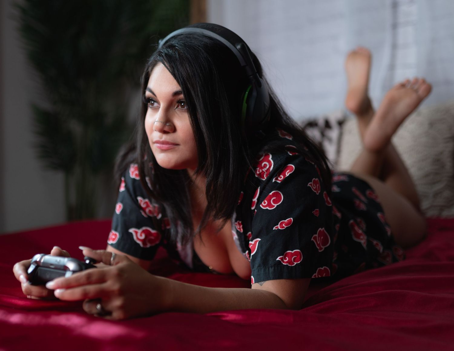 Young woman with headphones lying on red bed holding game controller wearing Akatsuki print pajamas.