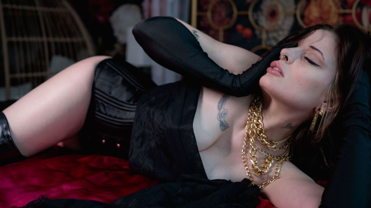 A person wearing dark clothing lies back with eyes closed and gold jewelry against a dramatic red background.