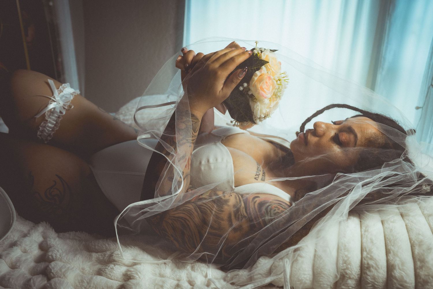 Tattooed bride in white lingerie and veil lying down holding a floral bouquet in a dreamy boudoir setting.