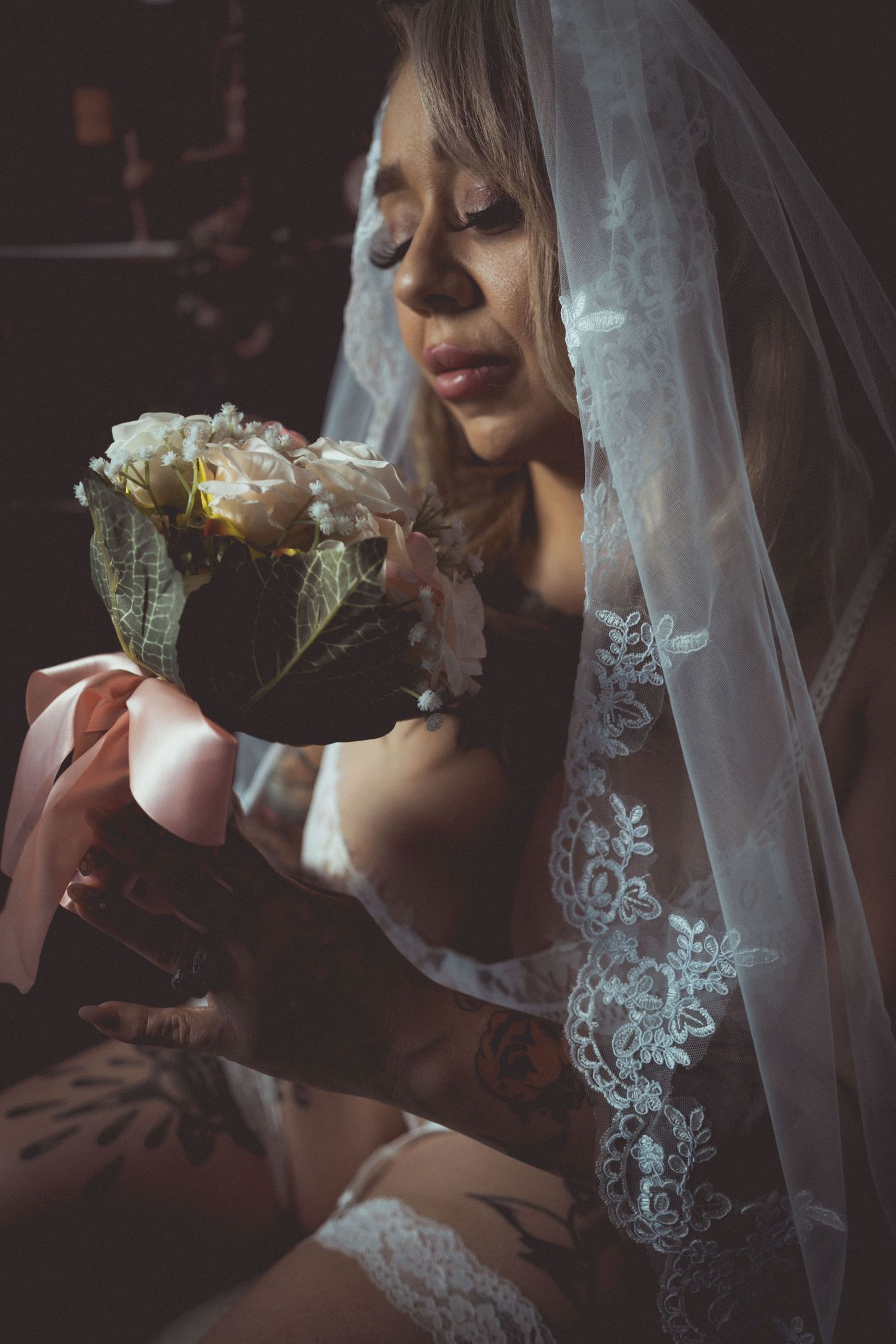 Tattooed bride in lace veil and lingerie holding a white rose bouquet in a dark, moody boudoir setting.