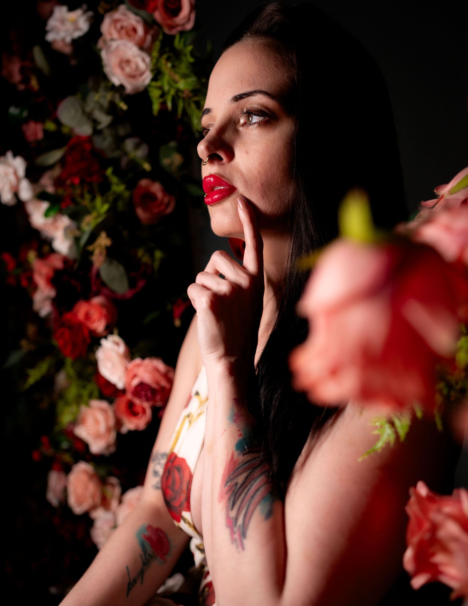 Woman with red lips and tattoos poses thoughtfully among pink and red roses in a dark, moody floral setting.
