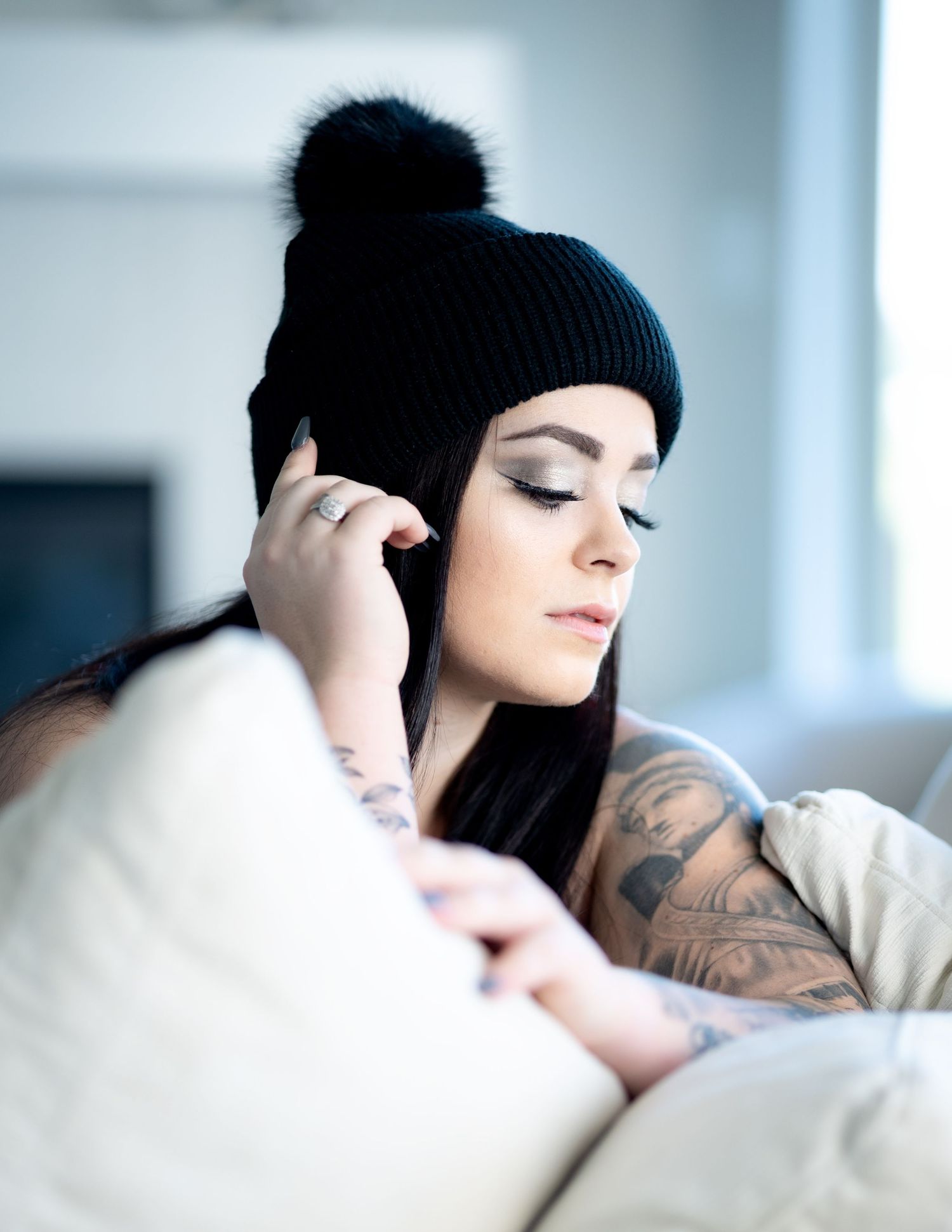 Person wearing black beanie and tattoos relaxes on white couch while using phone in soft natural light.