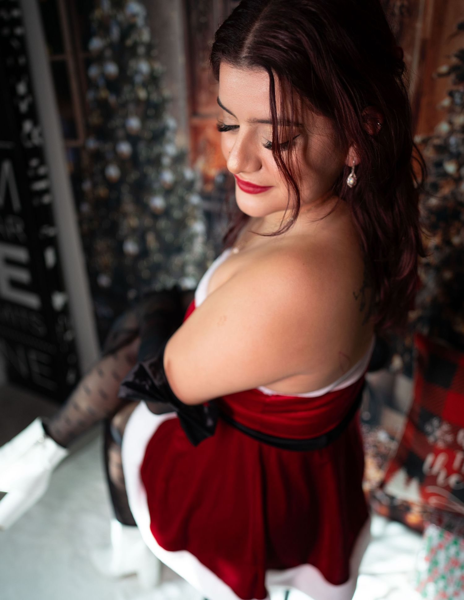 Woman in red velvet Santa dress with black gloves poses near decorated Christmas tree holiday backdrop.
