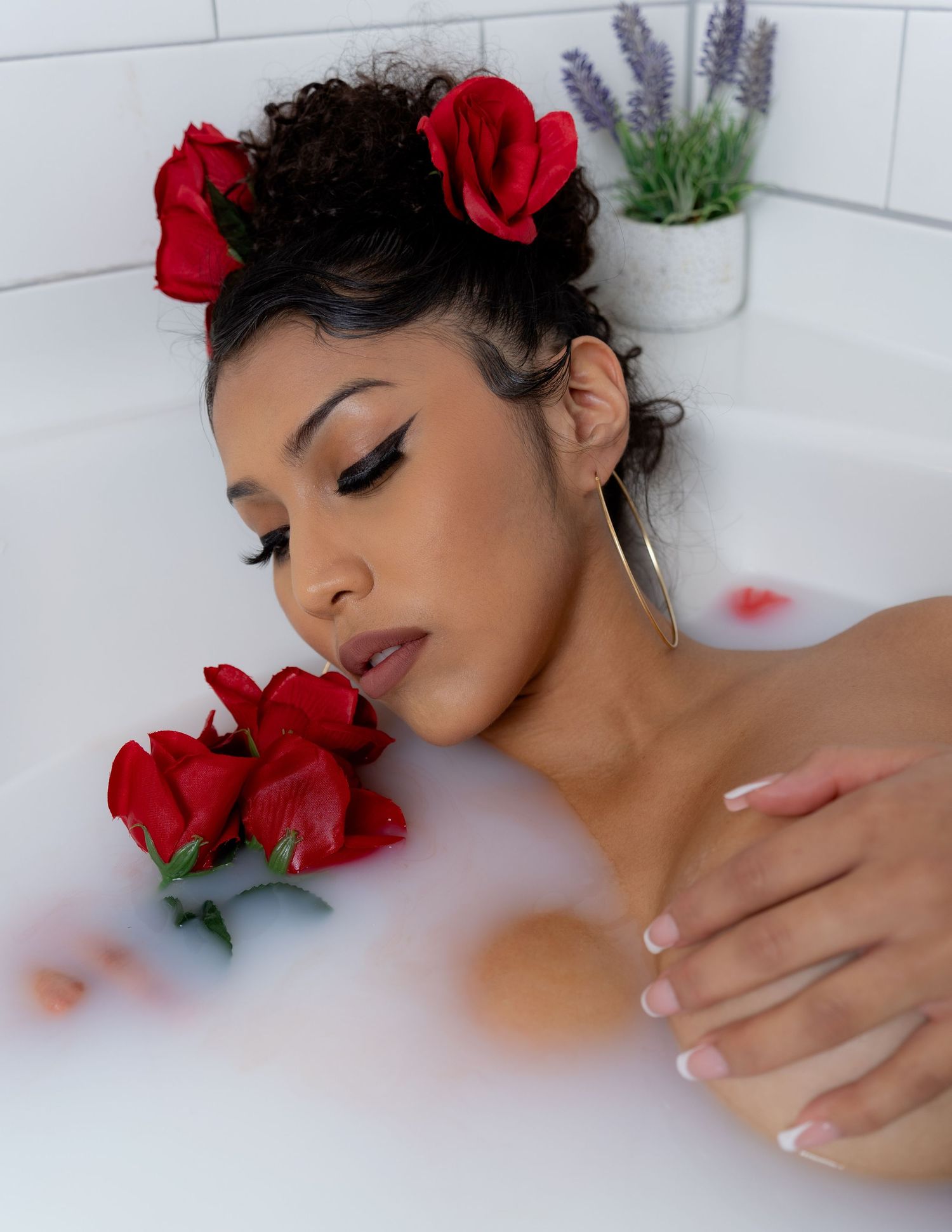 Woman with red roses in hair relaxing in a milky floral bath, wearing hoop earrings with elegant makeup.