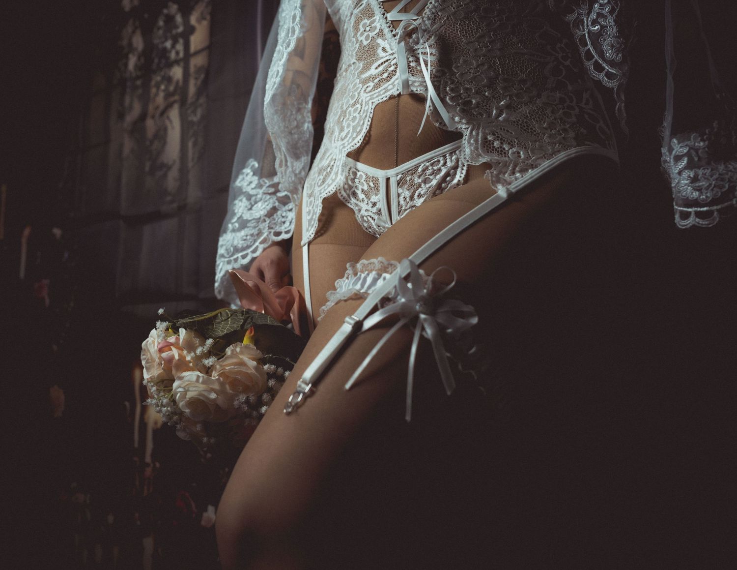 Bride in white lace lingerie and veil holding a bouquet, wearing a garter on her thigh.