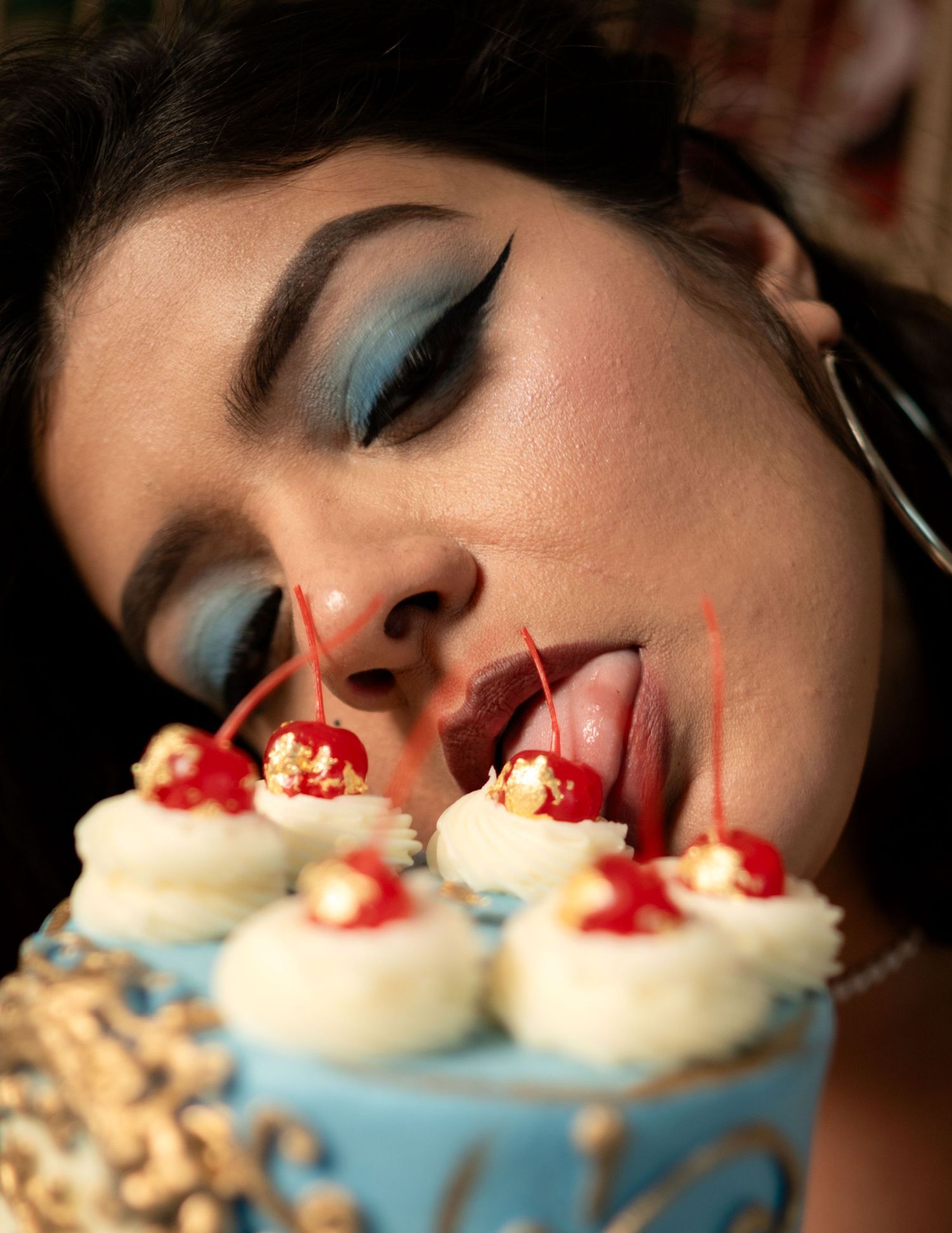 Woman with blue eyeshadow and dramatic eyeliner licking cherries off a decorative blue birthday cake.