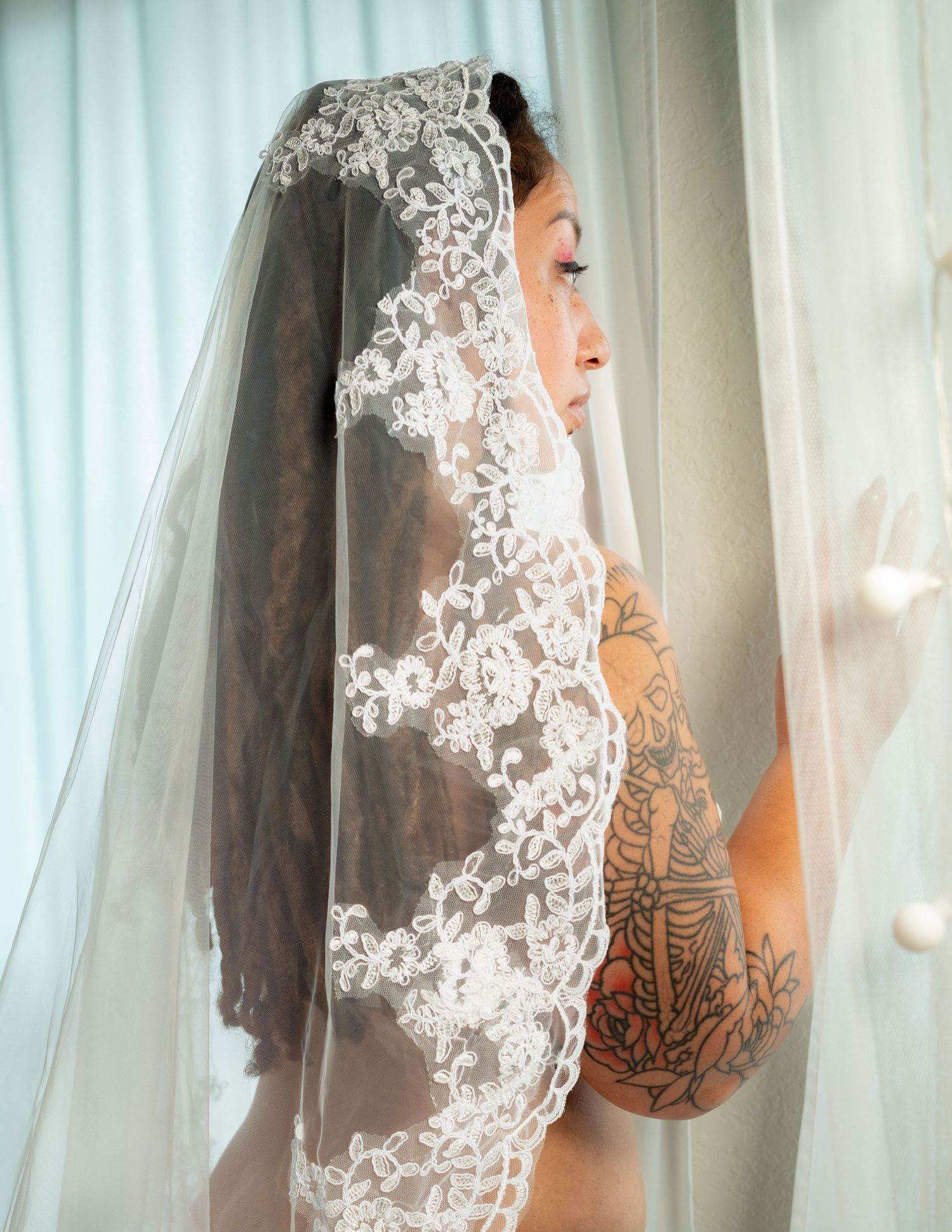 Bride wearing an intricate lace wedding veil with floral embroidery, showcasing tattoos on her shoulder and back.