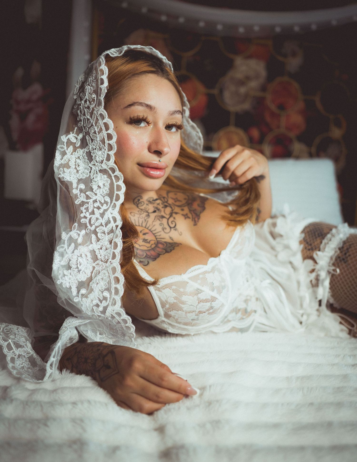 Woman in white lace lingerie and veil with tattoos posing elegantly on a bed in a boudoir setting.