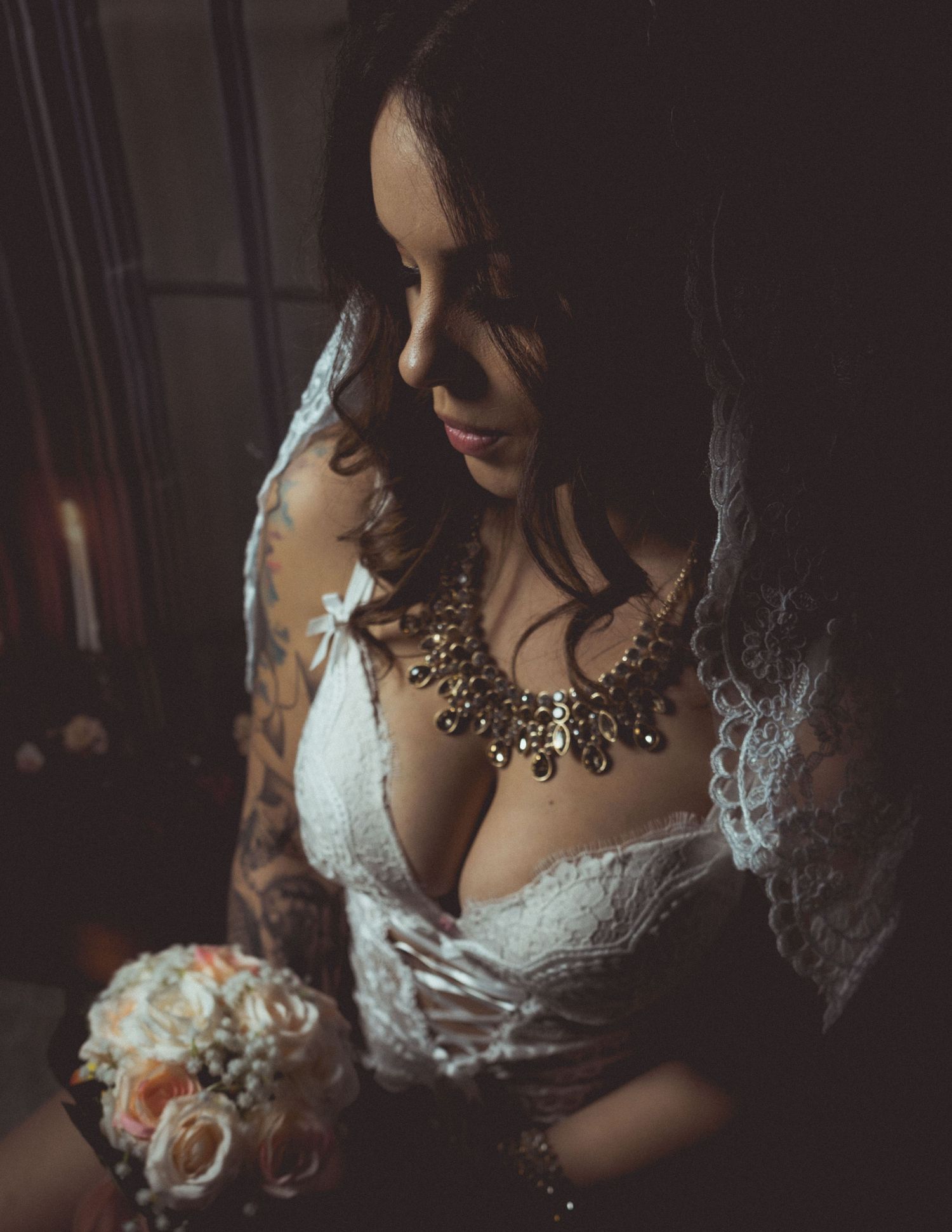 Bride in white lace gown with statement necklace holding a bouquet, tattoos visible on arms.