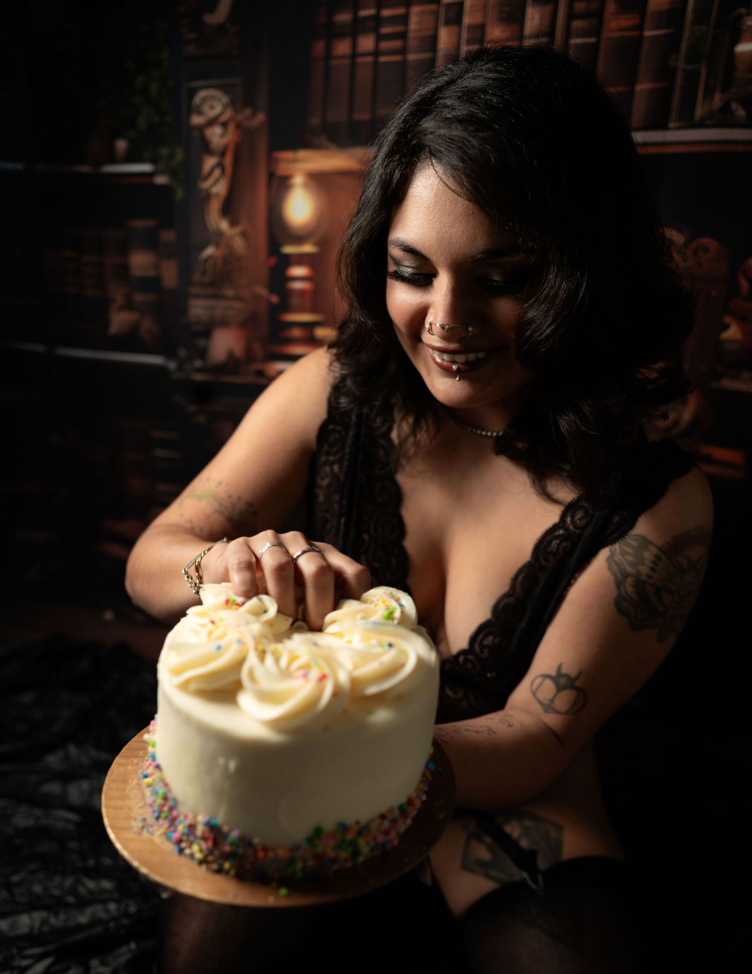 Woman with tattoos smiling while touching a white frosted celebration cake with sprinkles in a moody library setting.