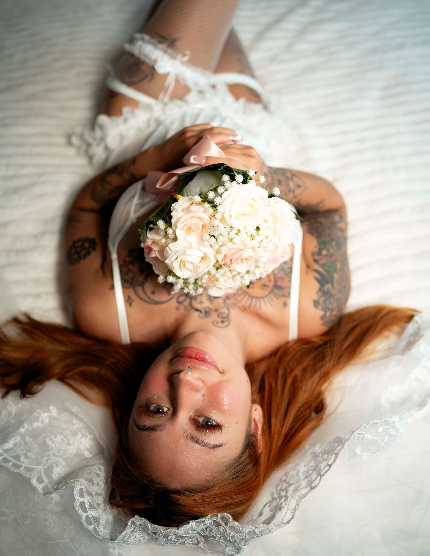 Tattooed redhead bride lying down holding a white and pink rose bouquet in white lace lingerie.