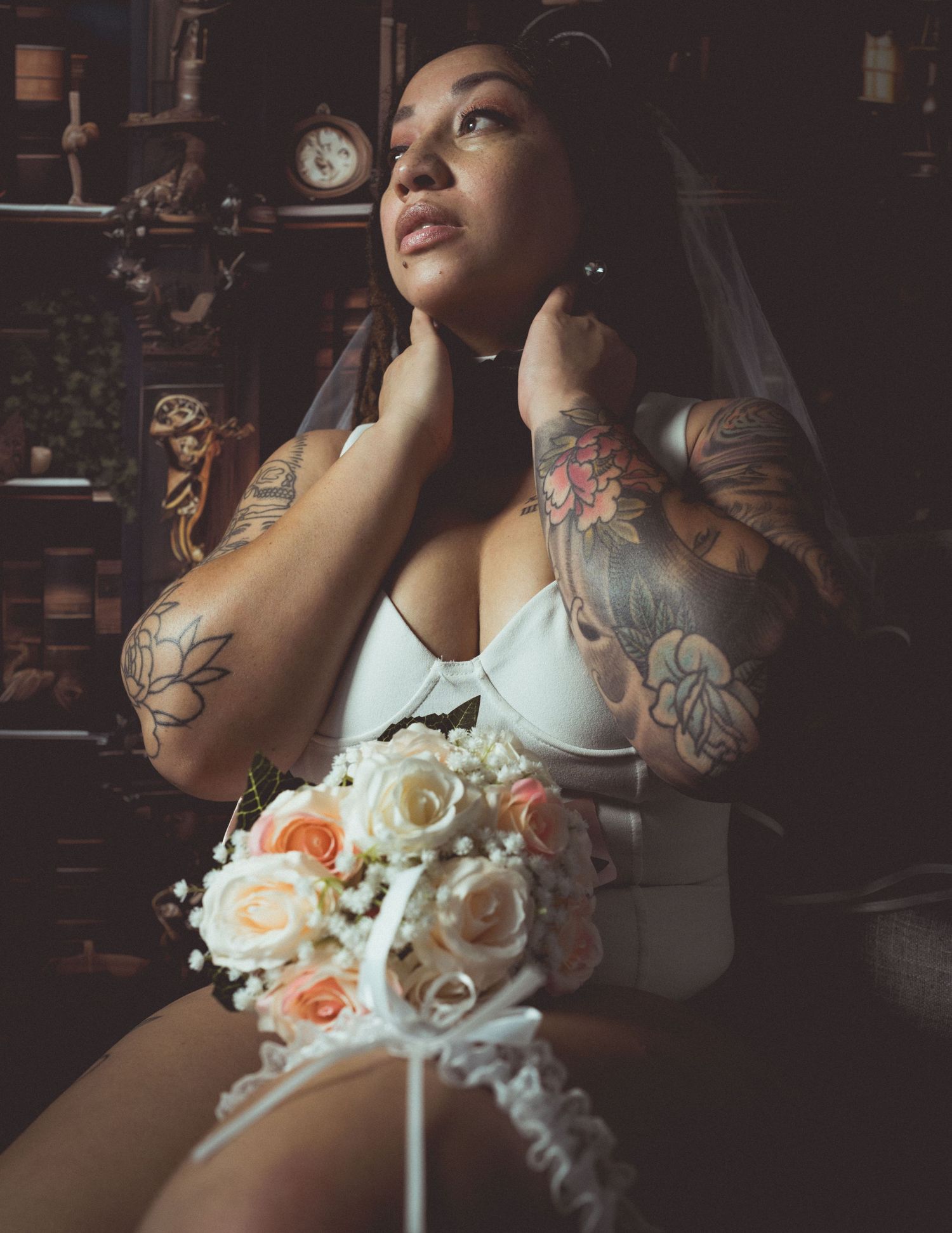 Tattooed bride in white lingerie and veil holds a bouquet of roses in a vintage-styled room.