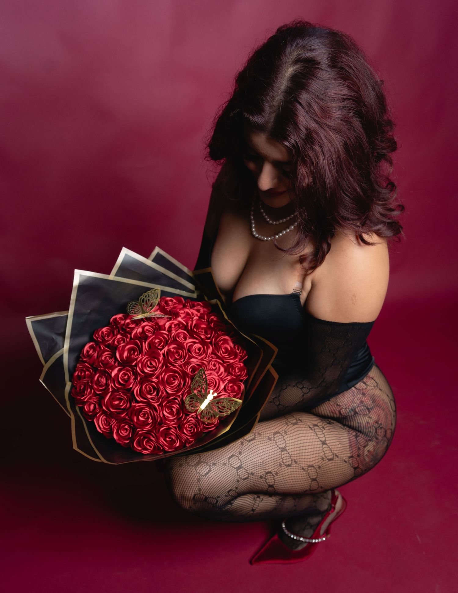 Woman in black lingerie sitting gracefully holding a large bouquet of red roses wrapped in black paper.