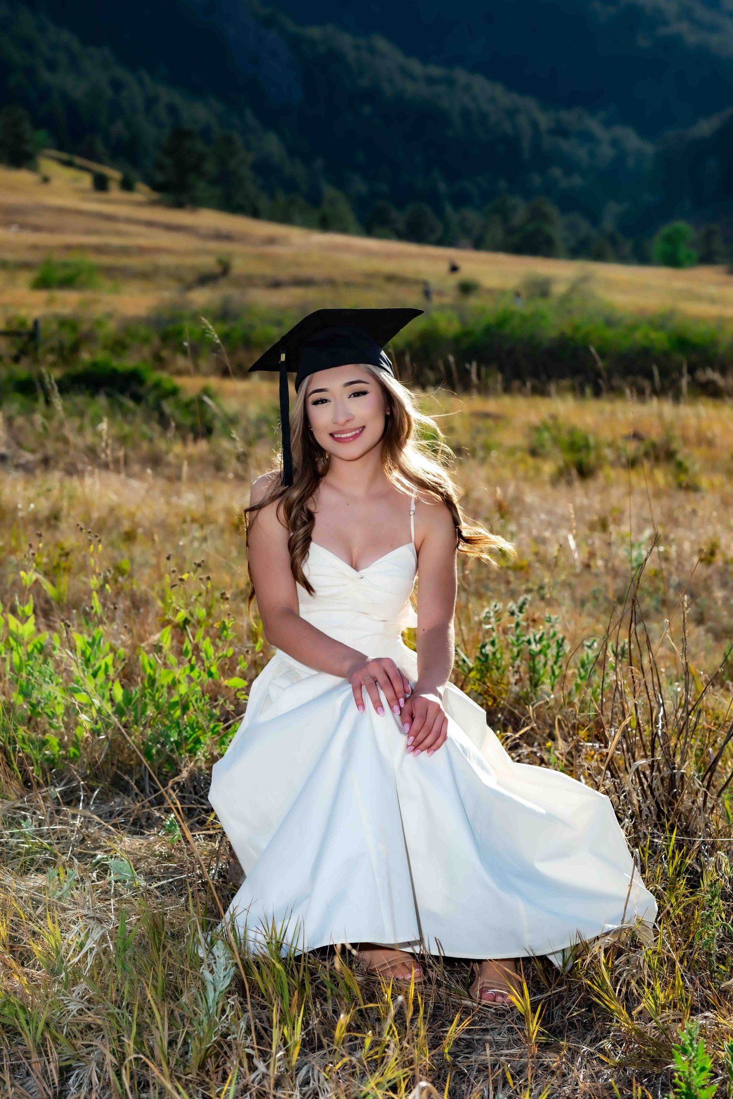 Smiling graduate in white dress and cap sits in a sunlit mountain meadow for senior portrait.