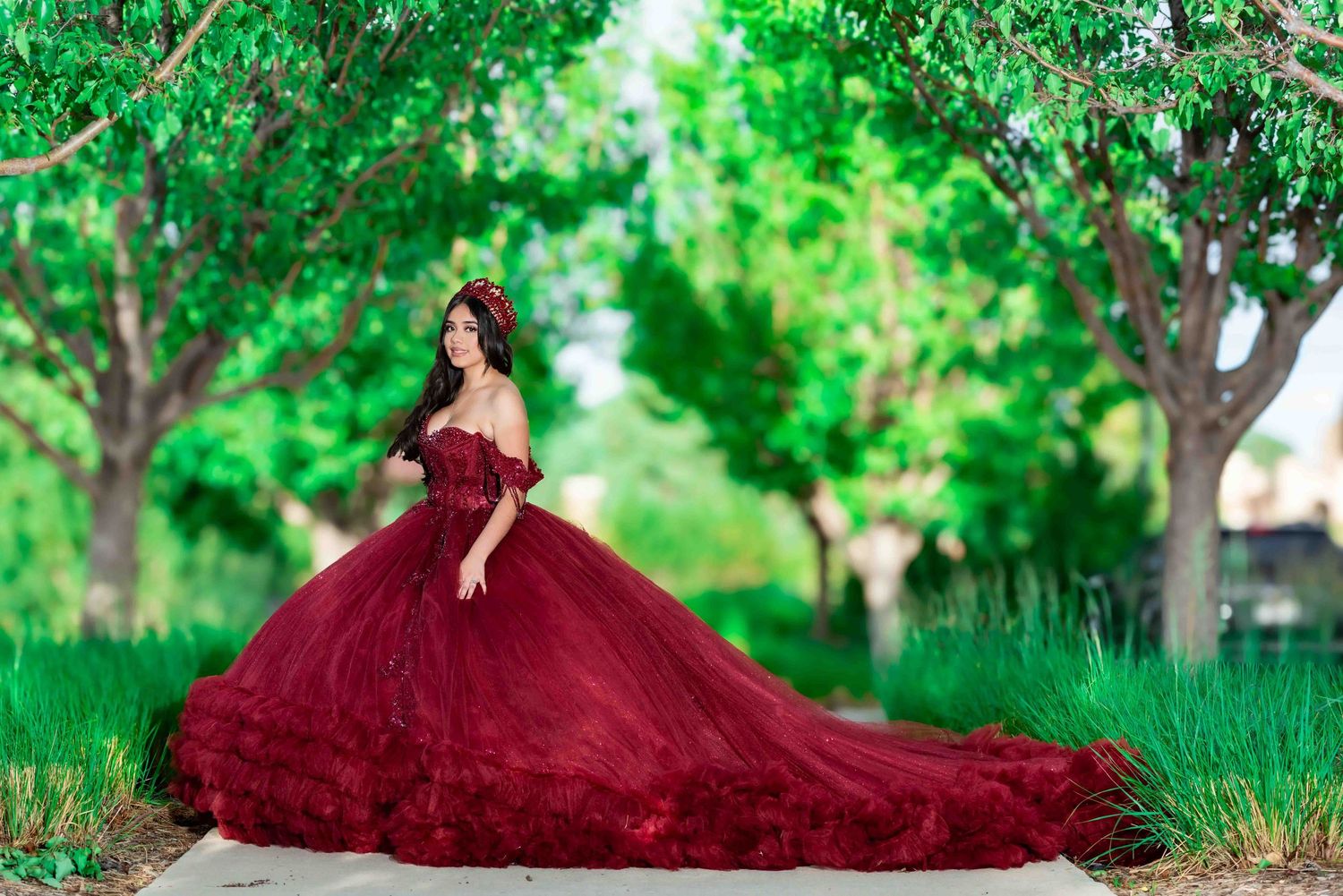 Young woman in voluminous dark red quinceañera ball gown poses gracefully along a tree-lined path.
