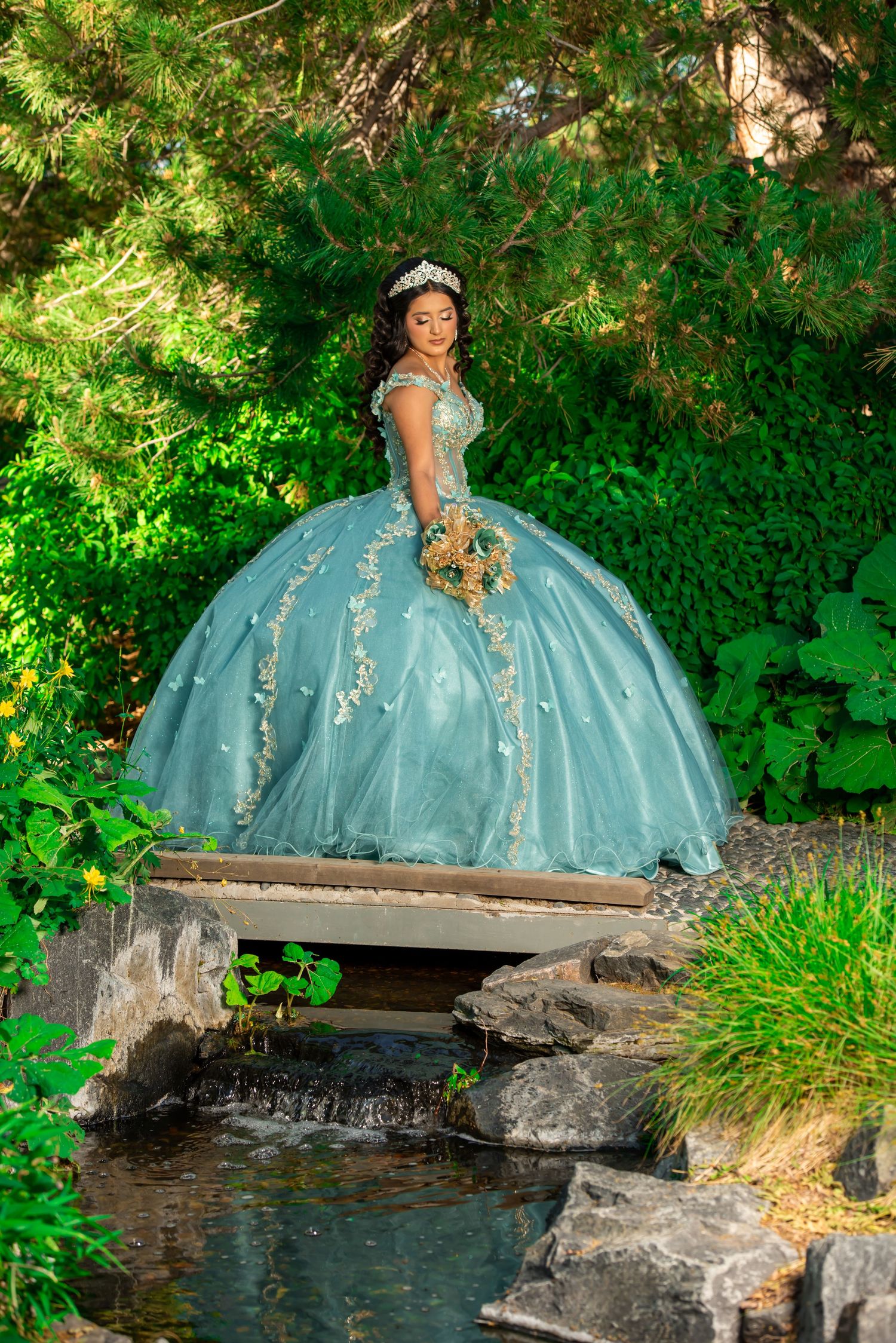 Quinceañera girl in elegant teal ball gown with floral embroidery stands on a garden bridge.