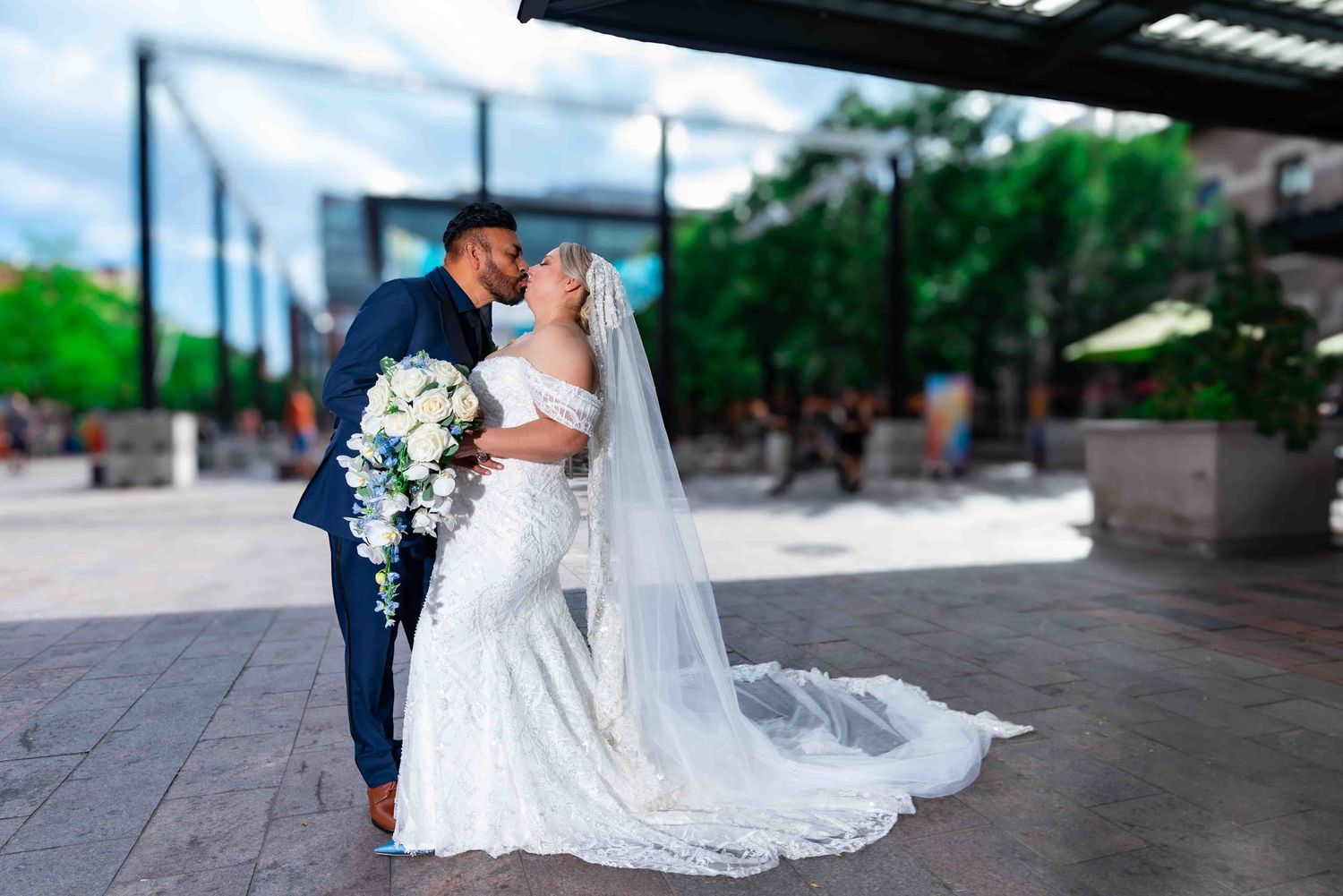 Bride and groom share a romantic kiss outdoors, bride wearing lace mermaid gown with long veil.