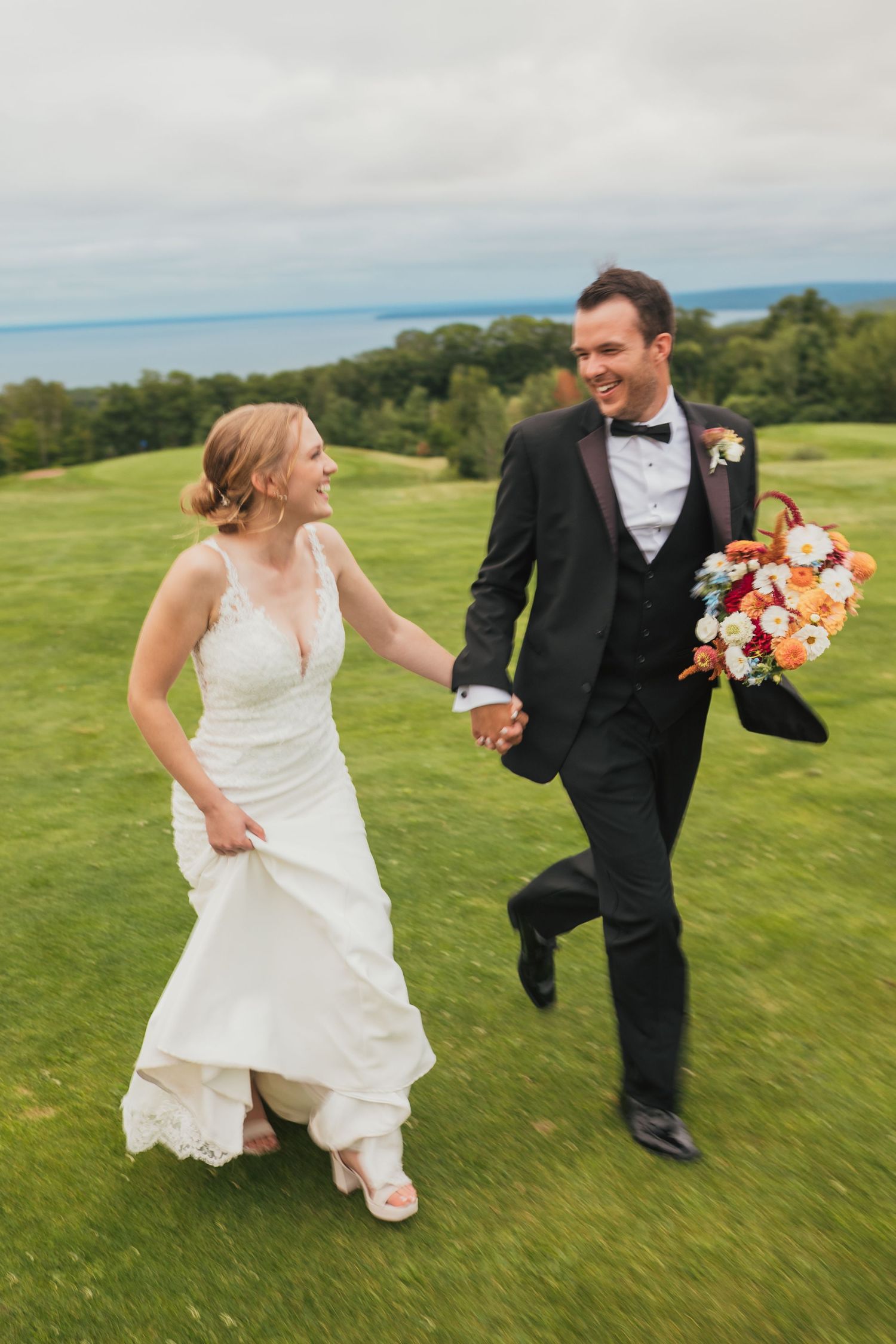 A playful couple in wedding attire runs and chases each other across a scenic hilltop meadow with rolling green hills in the background.