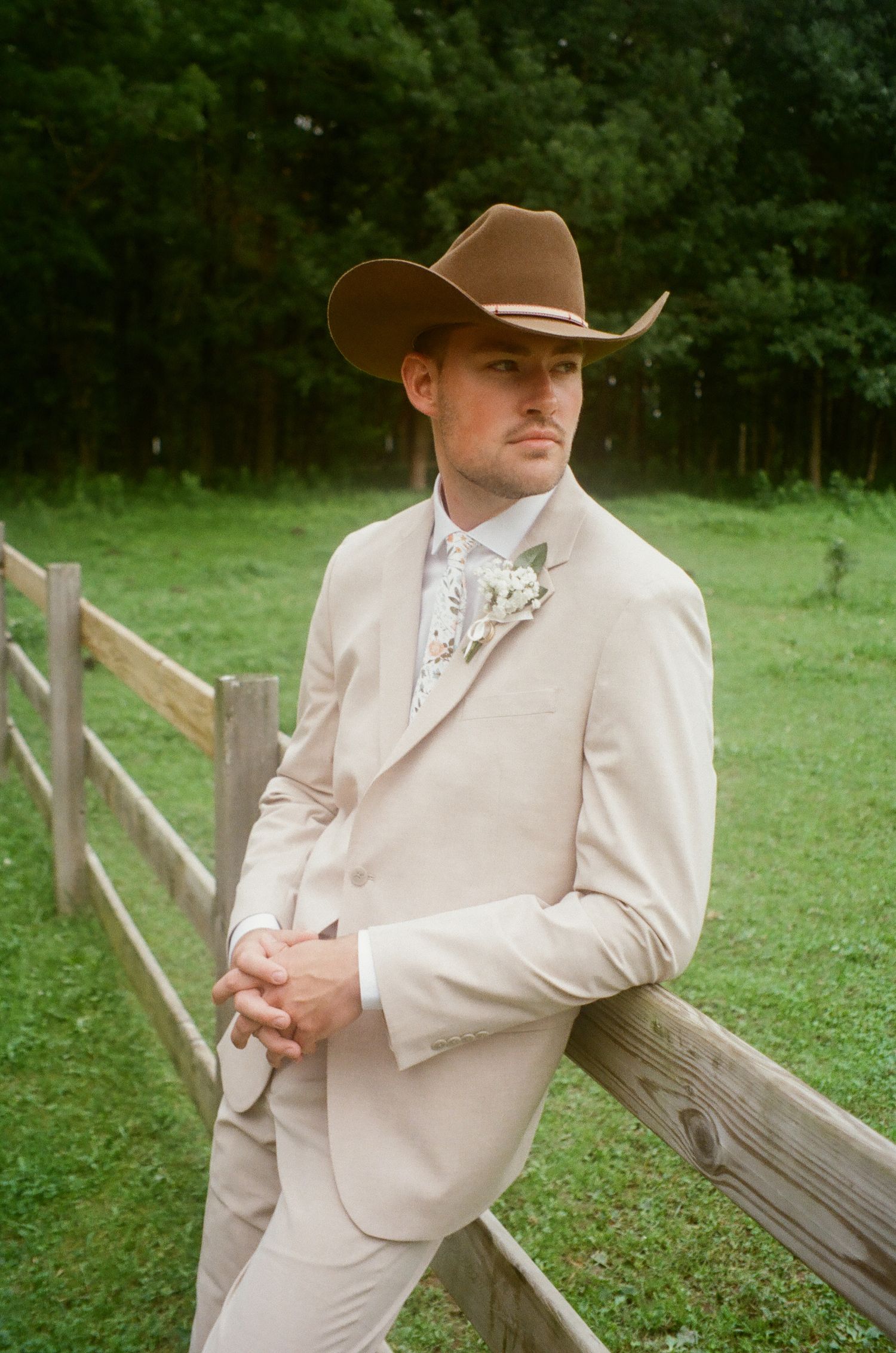 A person in a cream-colored suit and cowboy hat poses casually against a rustic wooden fence in a grassy outdoor setting.