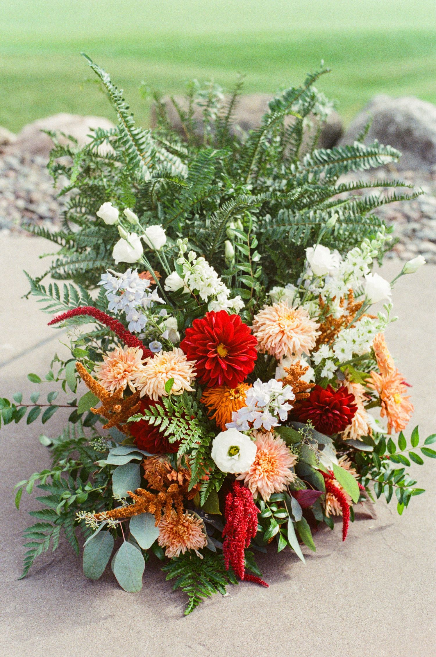 Floral arrangement with red dahlias, peach chrysanthemums, white roses, and green ferns displayed in a cascading style.