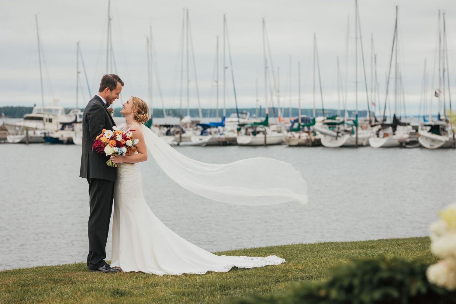 Newlyweds share a romantic kiss on a dock with sailboats and marina in the background.