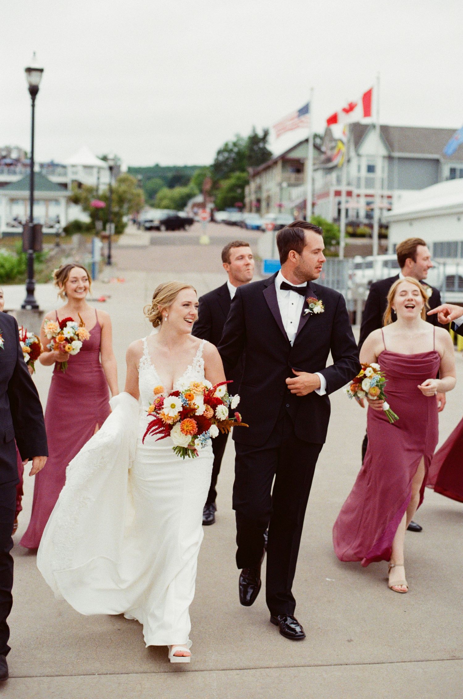 Wedding party walks and laughs together along waterfront street with American and Canadian flags in background.