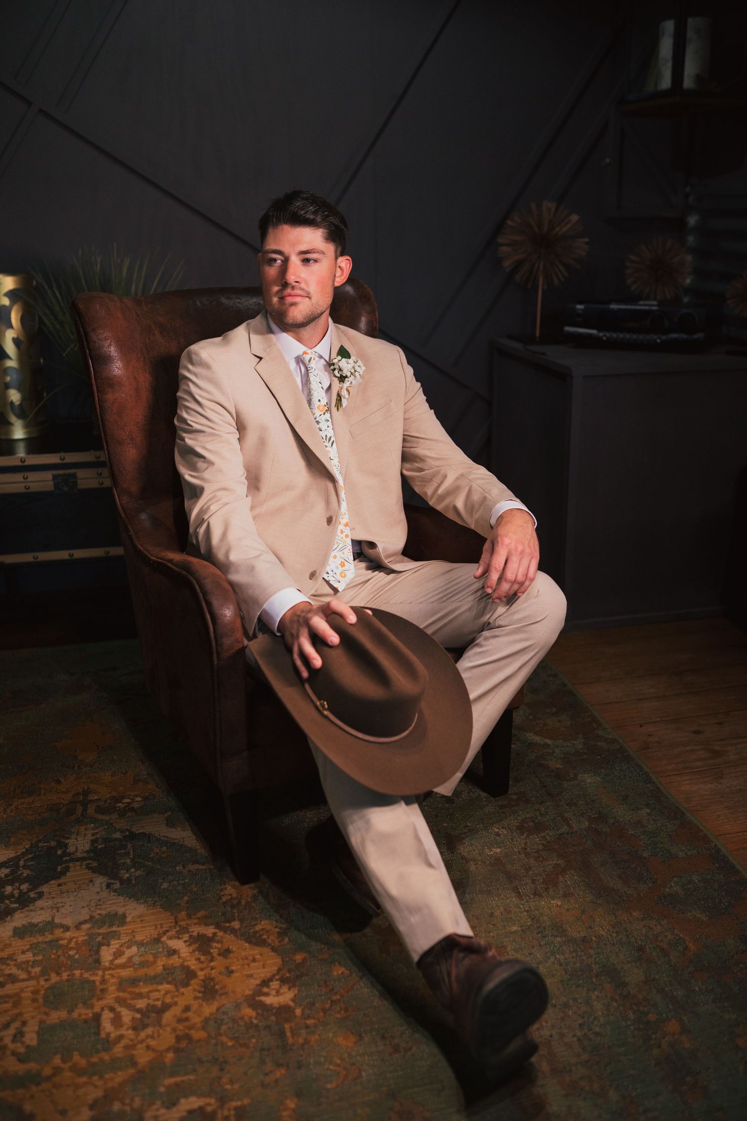 A person in a beige suit reclines in a brown leather chair on a patterned rug in a dimly lit room.