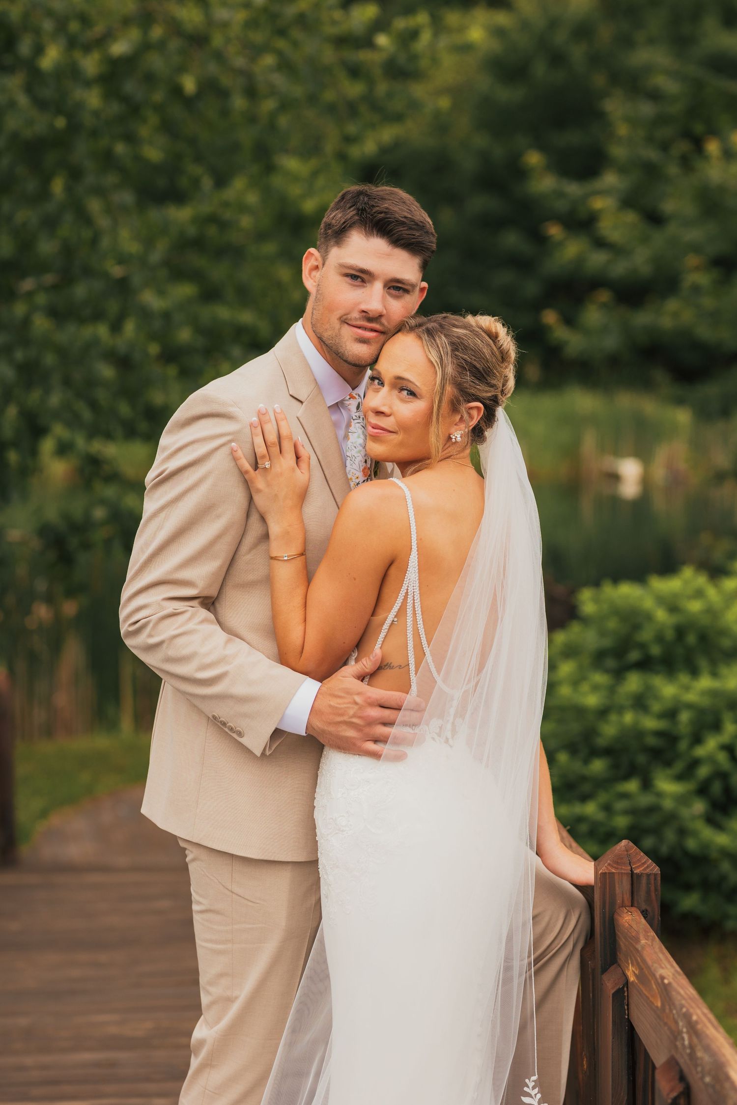 A couple in wedding attire share intimate moments on a wooden deck surrounded by lush greenery.