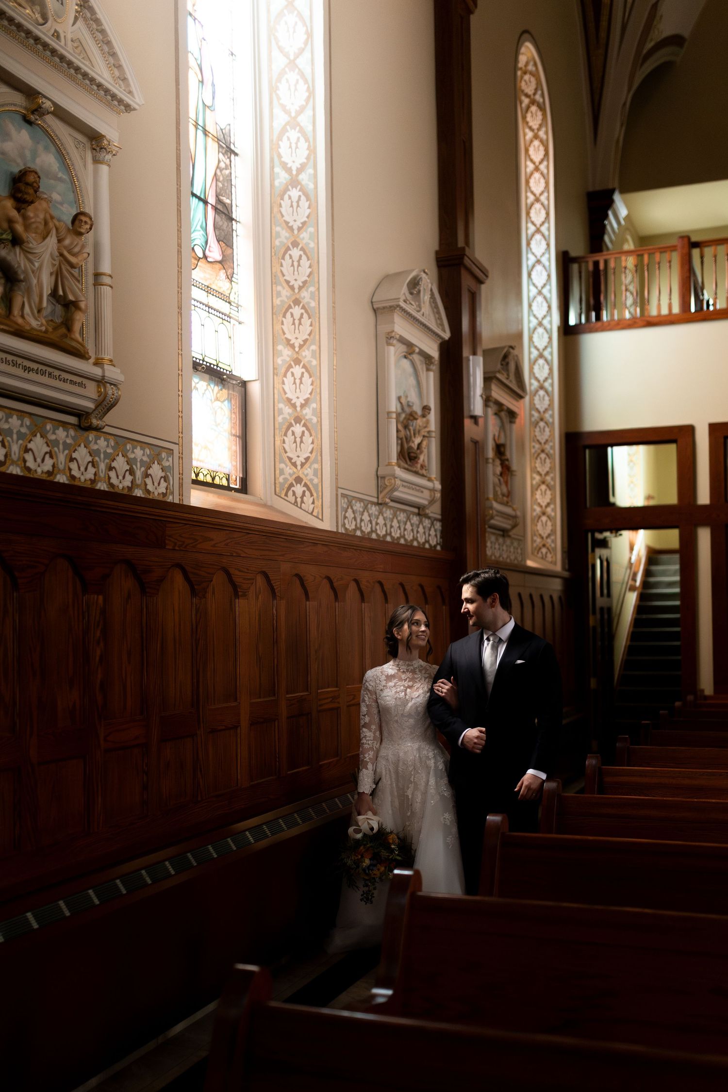 A couple stands together in the ornate wooden pews of a historic church with stained glass windows and decorative details.