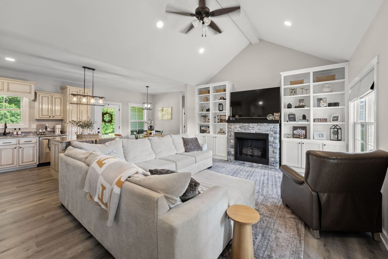 Spacious open-concept living room with vaulted ceiling, gray sectional sofa, built-in shelves, stone fireplace, and hardwood floors.
