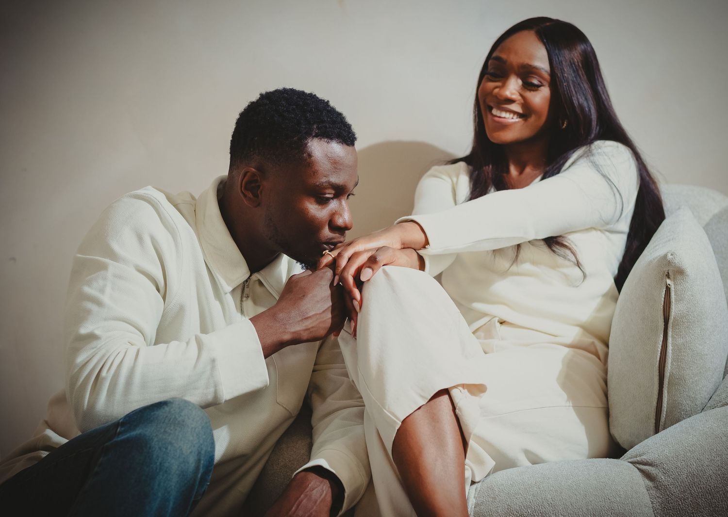 A man gently kisses his partner’s hand as she smiles, both dressed in light tones, capturing a moment of affection and connection.