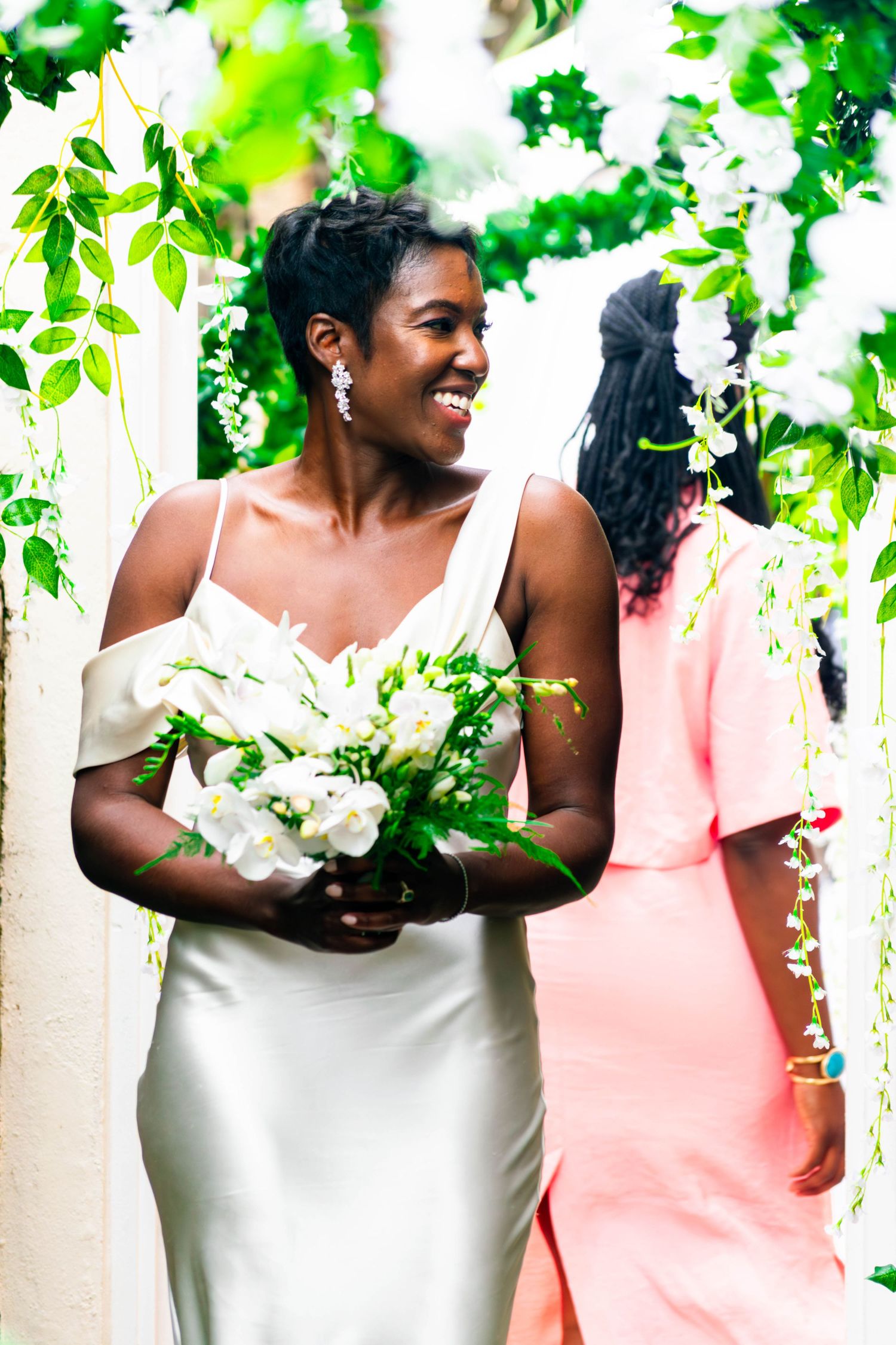 A joyful bride in an ivory gown holding a white floral bouquet, smiling as she walks through a lush green floral arch during her civil weddi