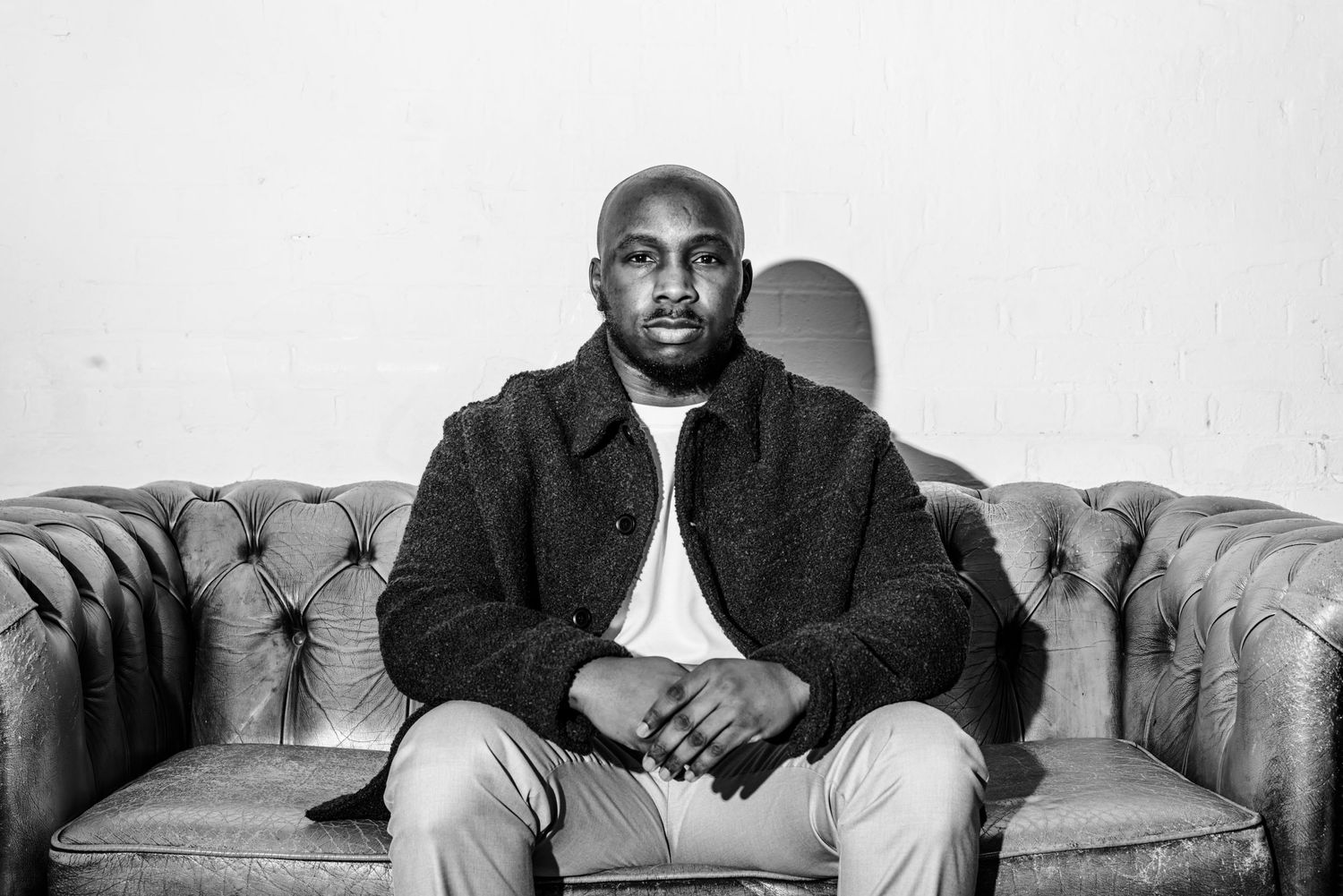 Black and white portrait of a man sitting on a leather sofa against a plain wall, hands folded, looking directly at the camera.