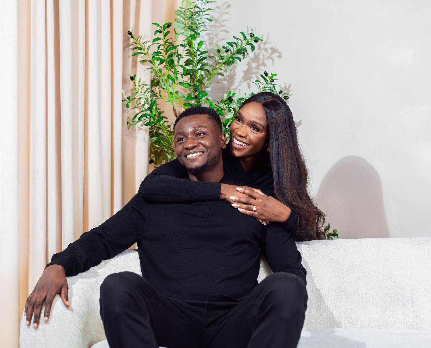 A smiling couple seated together on a cream sofa, the woman embracing her partner from behind, radiating warmth and joy.
