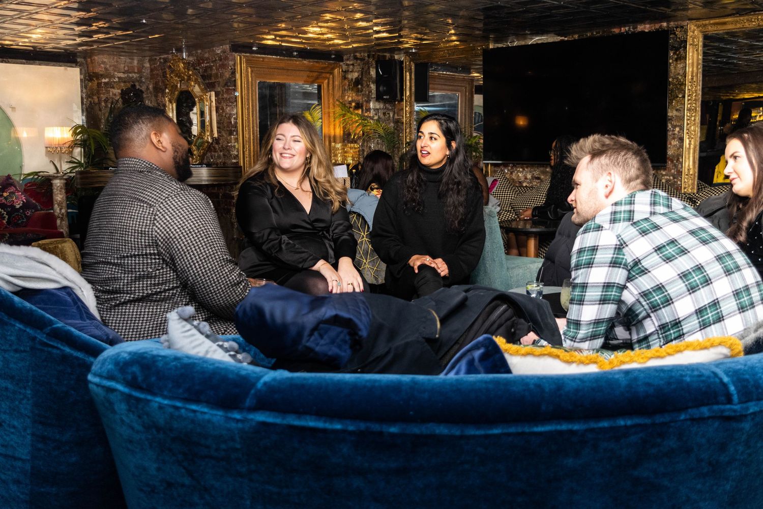 A small group of people sitting together on blue velvet sofas in a cozy lounge, engaged in lively conversation during an indoor event.