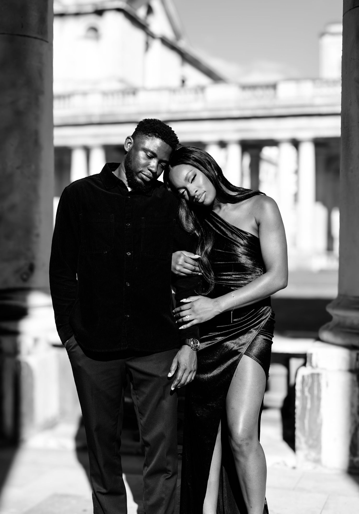 A couple stands close together in a black-and-white portrait; the woman rests her head on the man’s shoulder, capturing quiet intimacy.