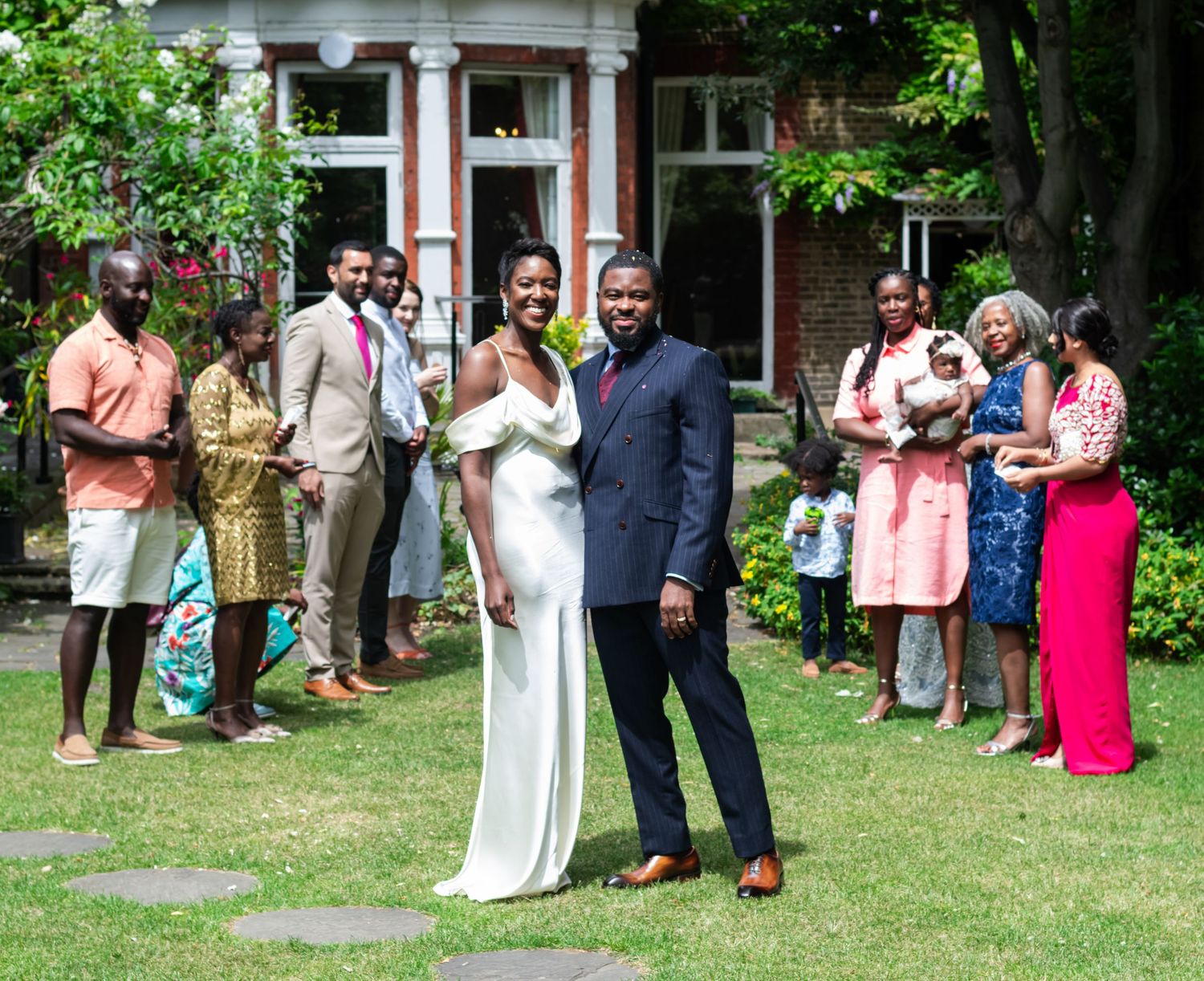A joyful group portrait taken outdoors after a civil wedding ceremony. The couple stands surrounded by family and friends dressed in bright,