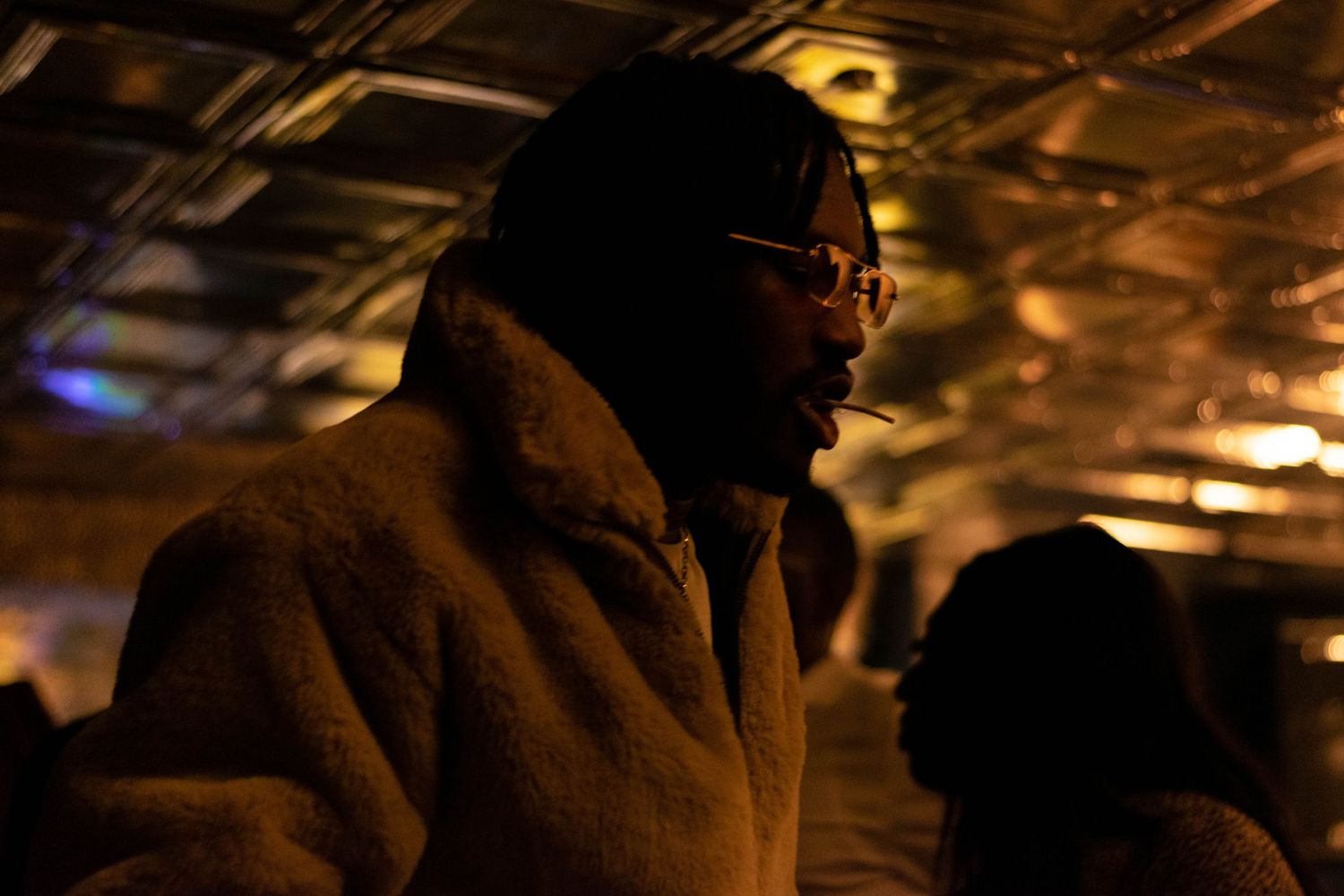 Man wearing a fur coat and sunglasses at a dimly lit event, captured in a candid moment