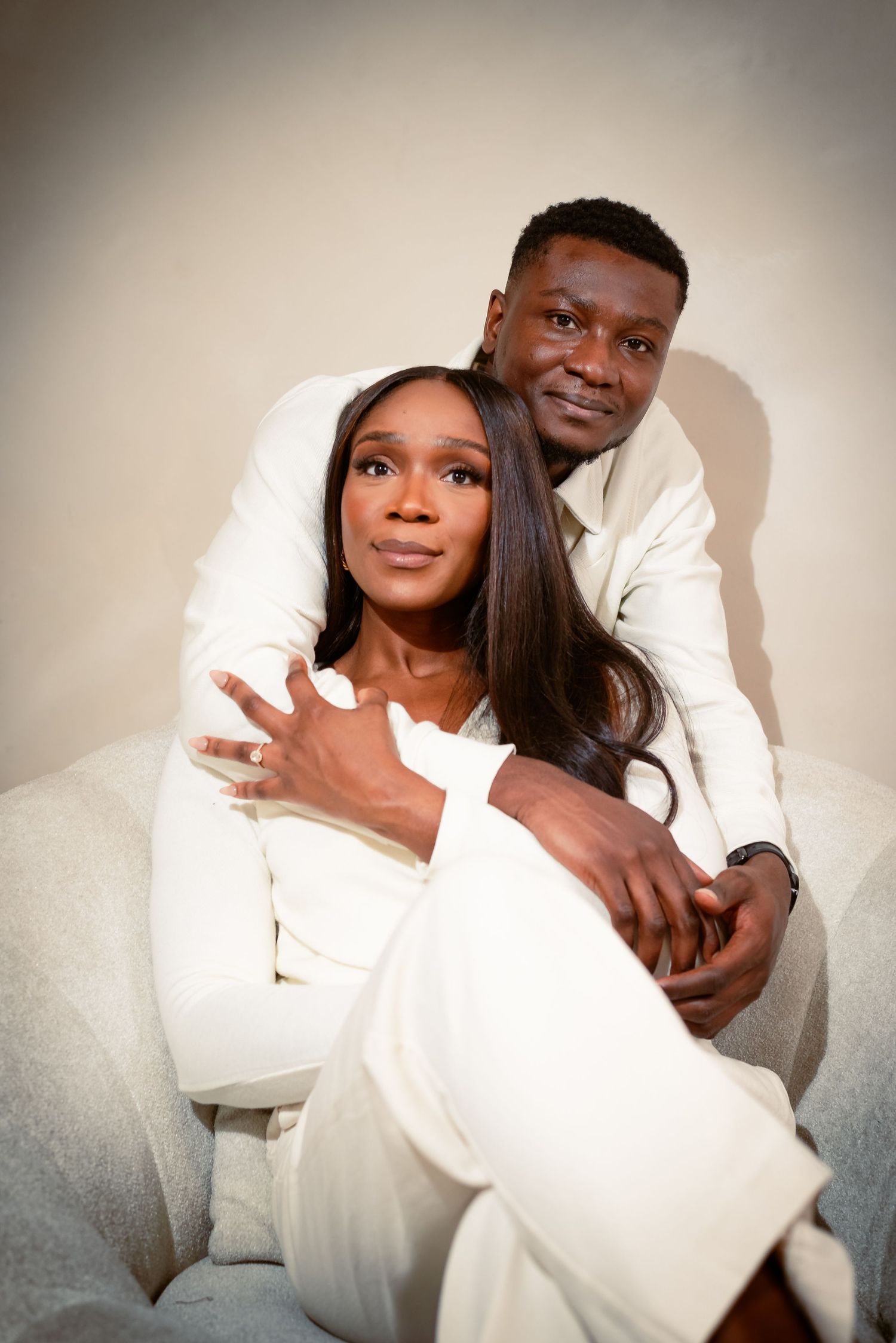 A couple dressed in soft cream tones sitting close together, sharing a tender embrace with calm expressions and warm intimacy.