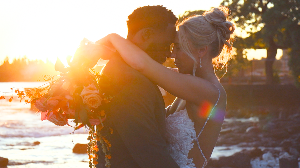 A bride holds a wedding boquet with her arms draped around the groom, with heads leaning and Kona, Hawai'i's sunset behind on the beach.