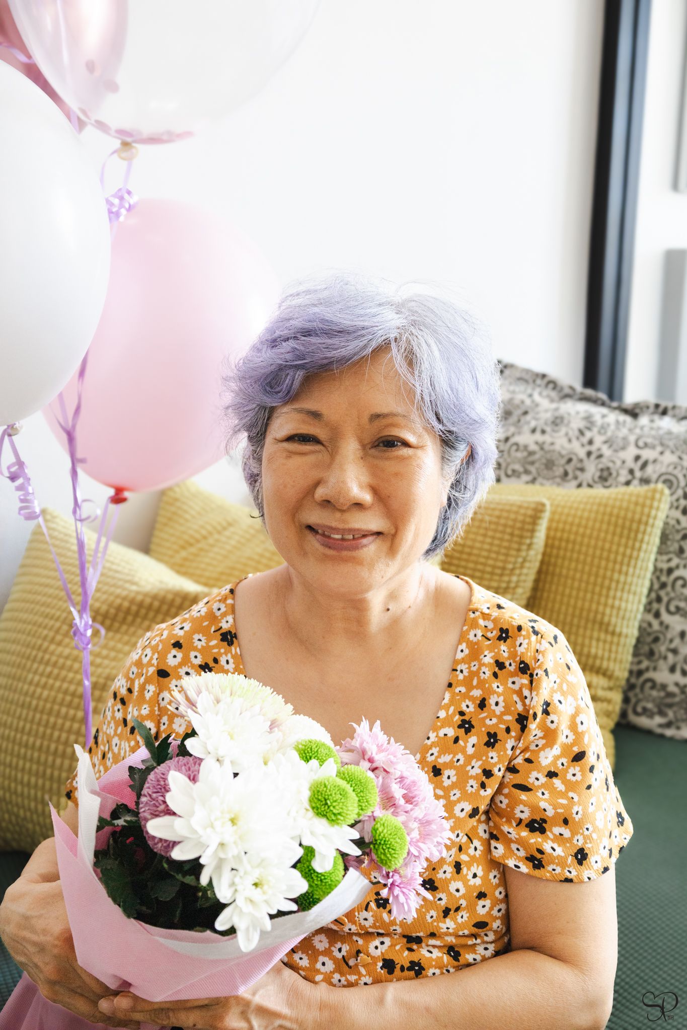 A photograph by Sara Pang (MSK Productions) of an elderly woman holding a bouquet of pink flowers and balloons behind her in Malaysia.