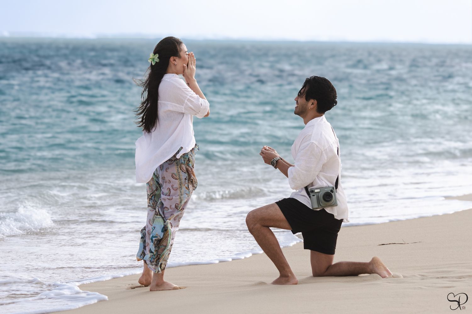 A fiancé kneels on Lanikai Beach on O'ahu as his fiancée covers her mouth with her hand in surprise.