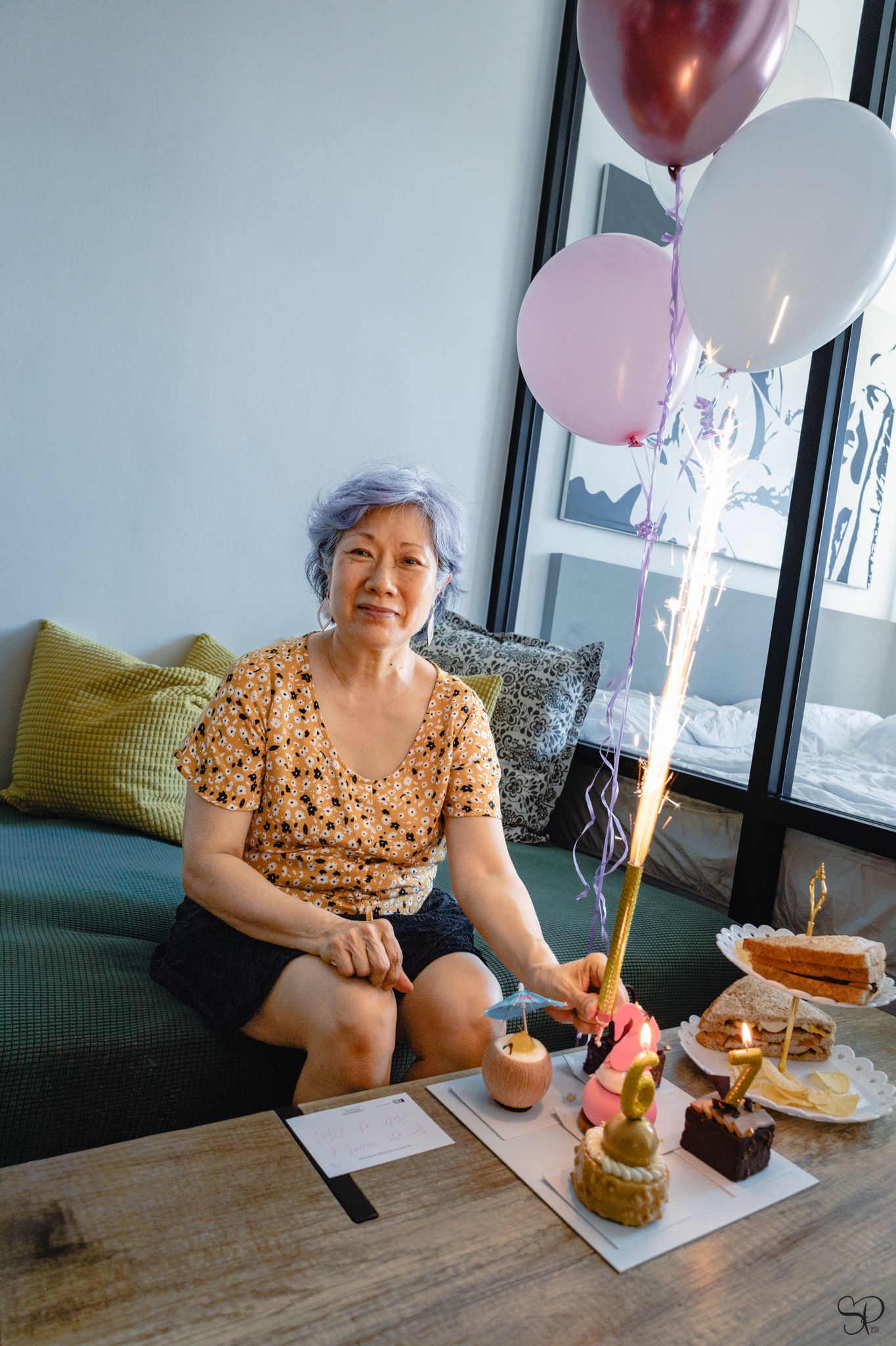 A photograph by Sara Pang (MSK Productions) of an elderly woman smiling with a sparkly birthday candle and balloons behind her in Malaysia.