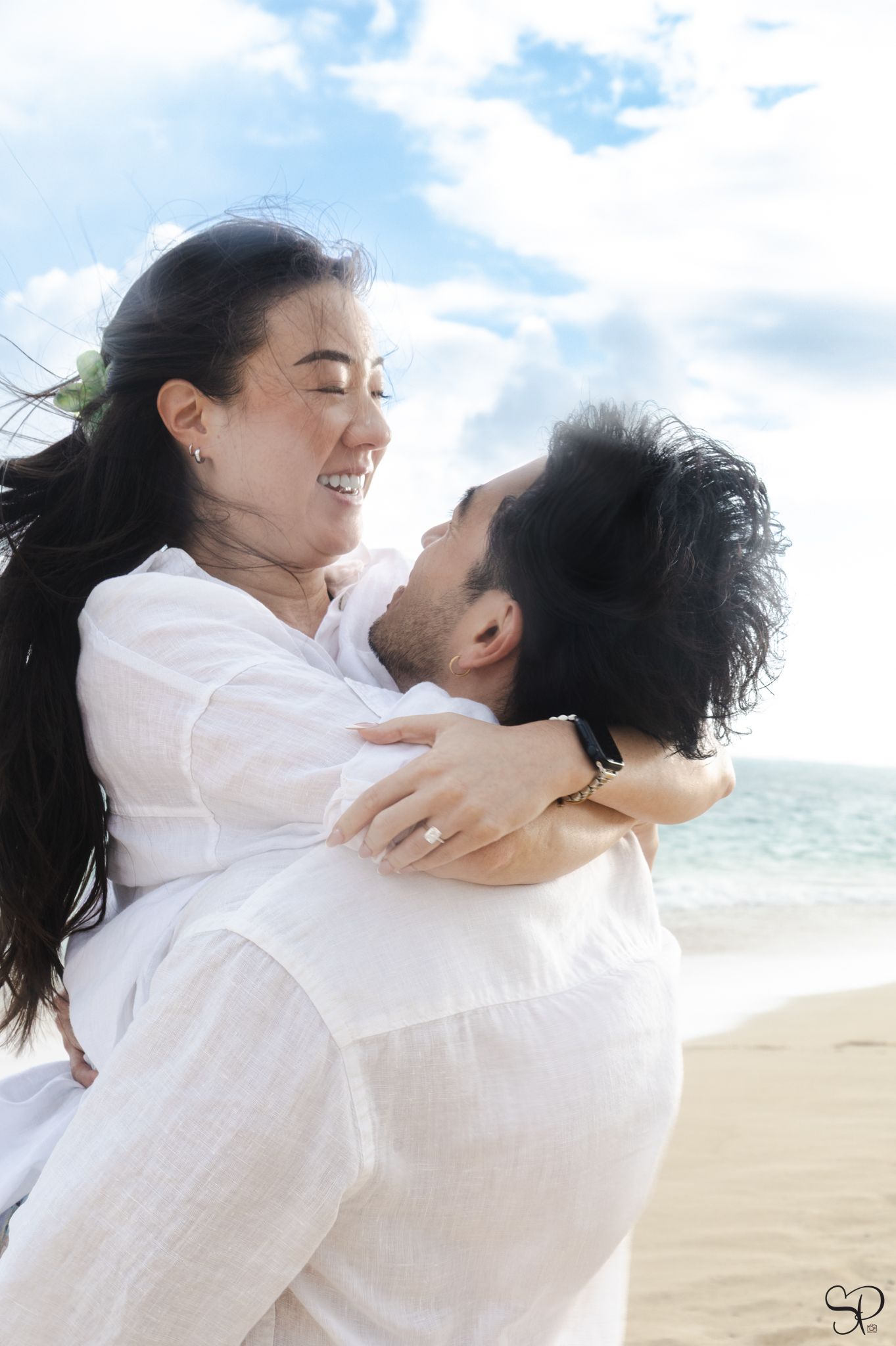 A fiancé lifts up his fiancée on Lanikai beach, O'ahu as she has her arms wrapped around him, smiling with hair blowing in the wind.