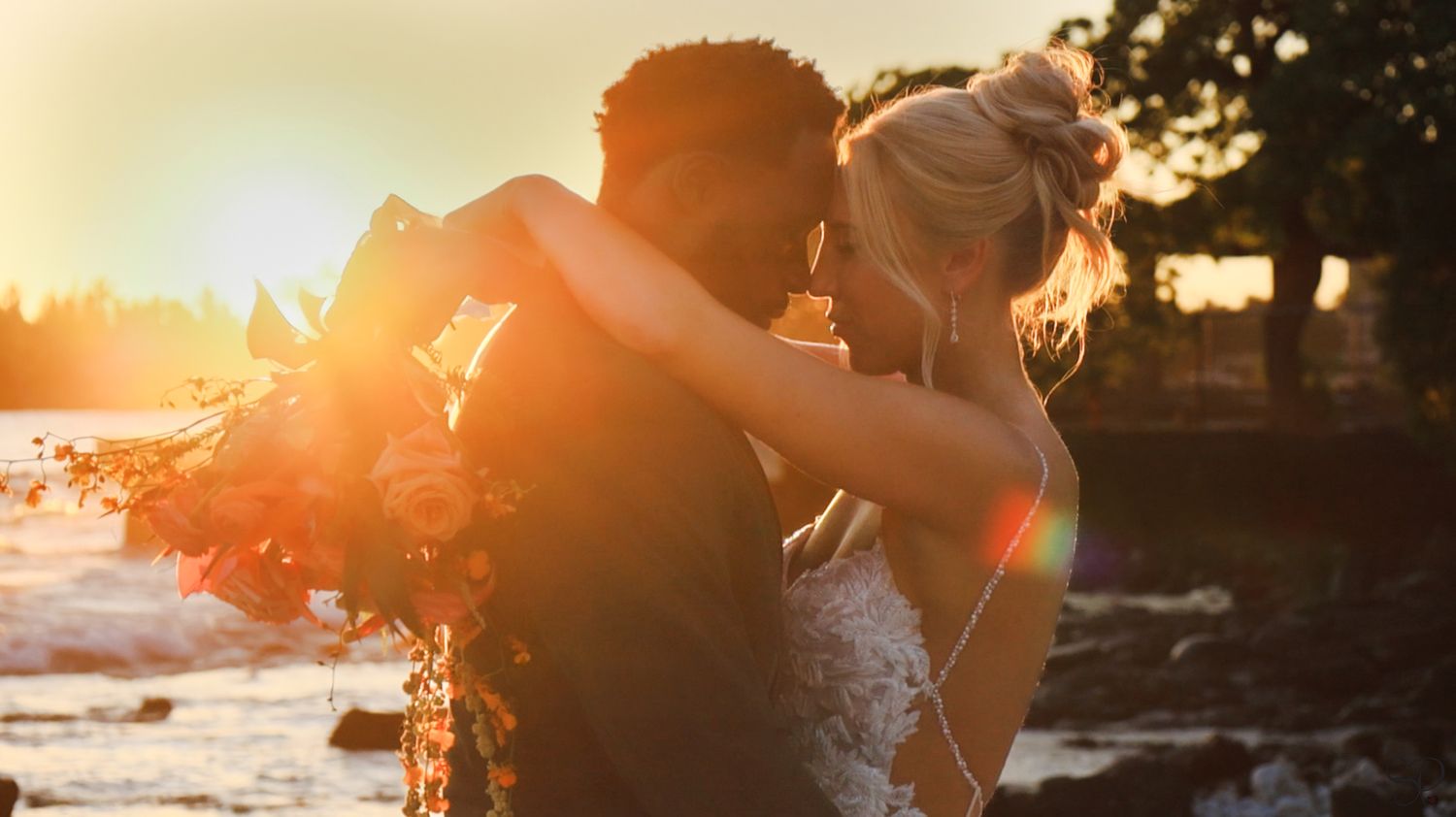 A bride holds a wedding boquet with her arms draped around the groom, with heads leaning and Kona, Hawai'i's sunset behind on the beach.