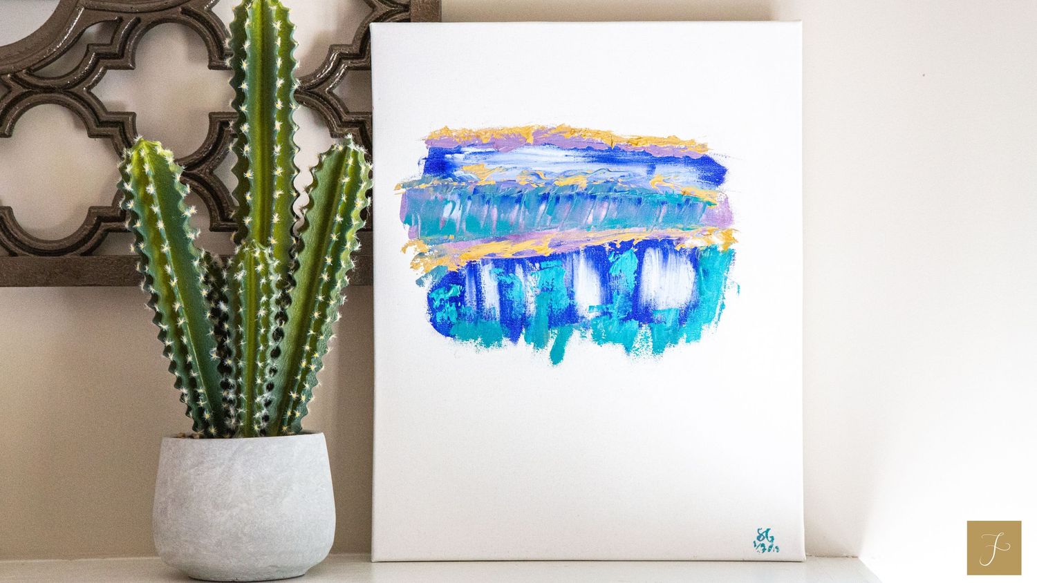 An abstract painting by Sara Pang with blue, gold, and green on the canvas sitting on a mantle with a green cactus plant beside it.